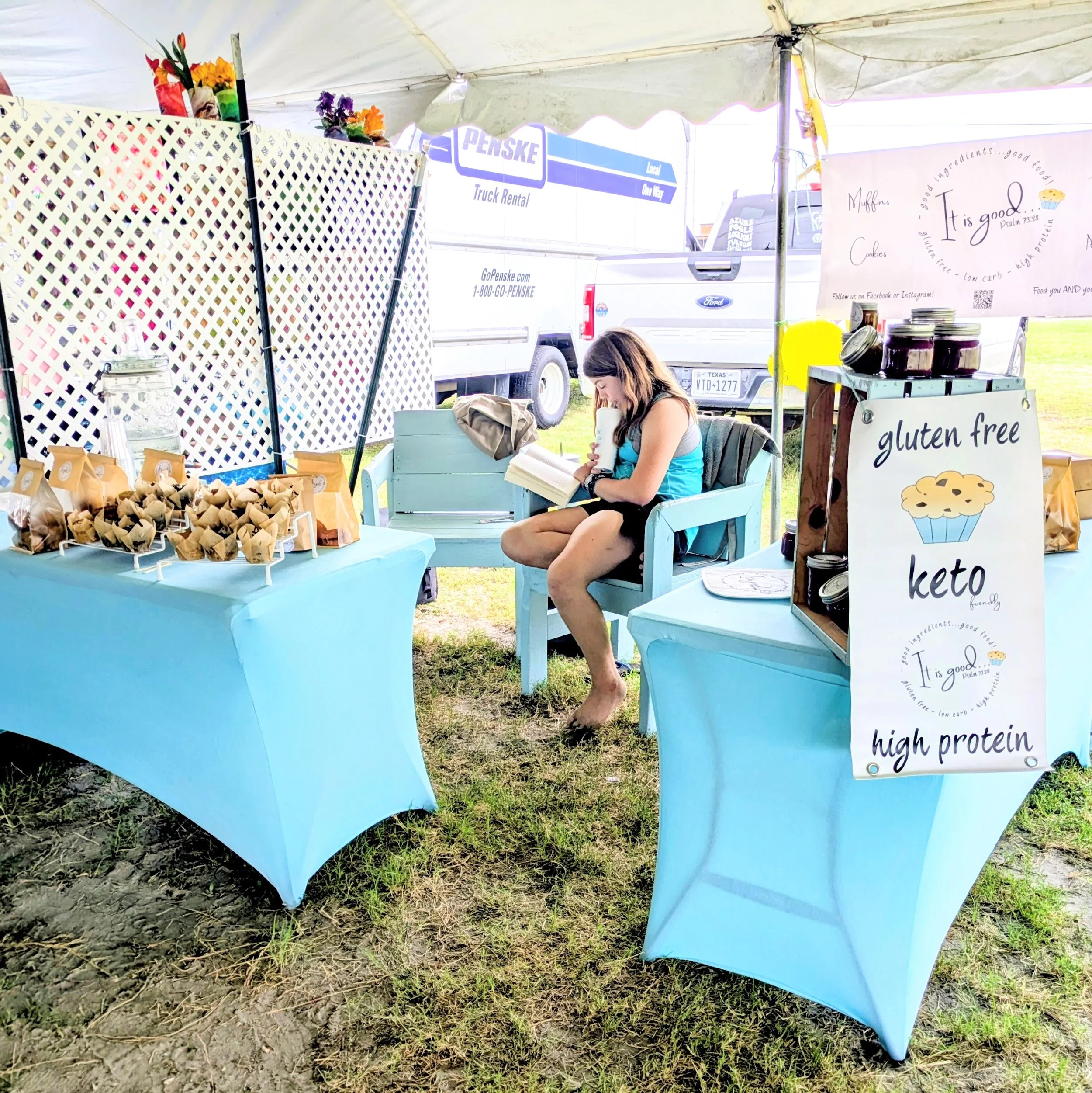 At an outdoor market stall, a woman sits on a light blue bench, reading a book and drinking from a mug. The stall has a blue tablecloth, various jars, and paper cones filled with baked goods. A sign advertises gluten-free, keto-friendly cookies and muffins, with mentions of high protein and low carb ingredients. There are colorful flowers and a white lattice divider, with a Penske truck visible in the background.