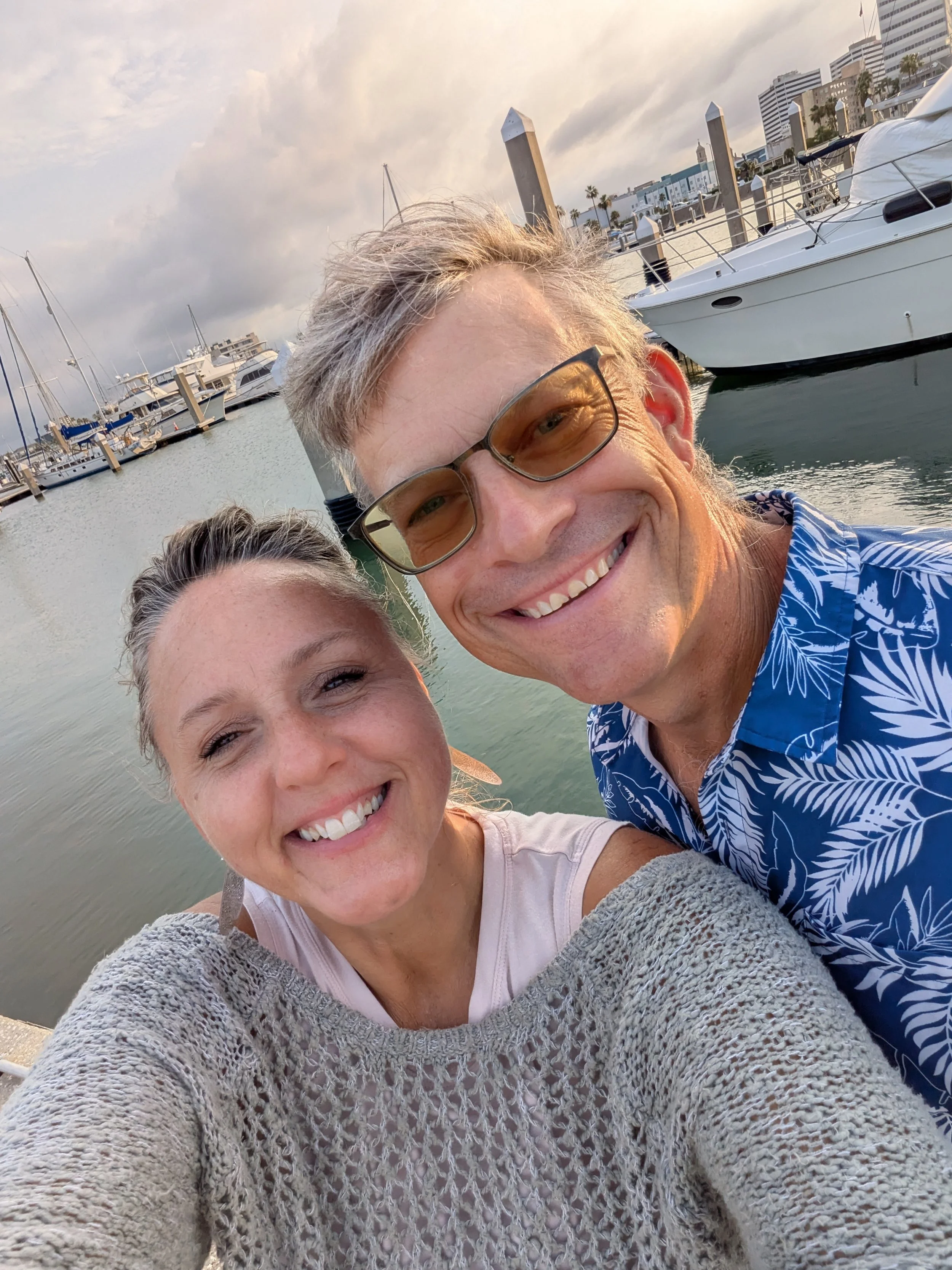 A smiling woman and a smiling man taking a selfie at a marina with boats docked in the background on a partly cloudy day.