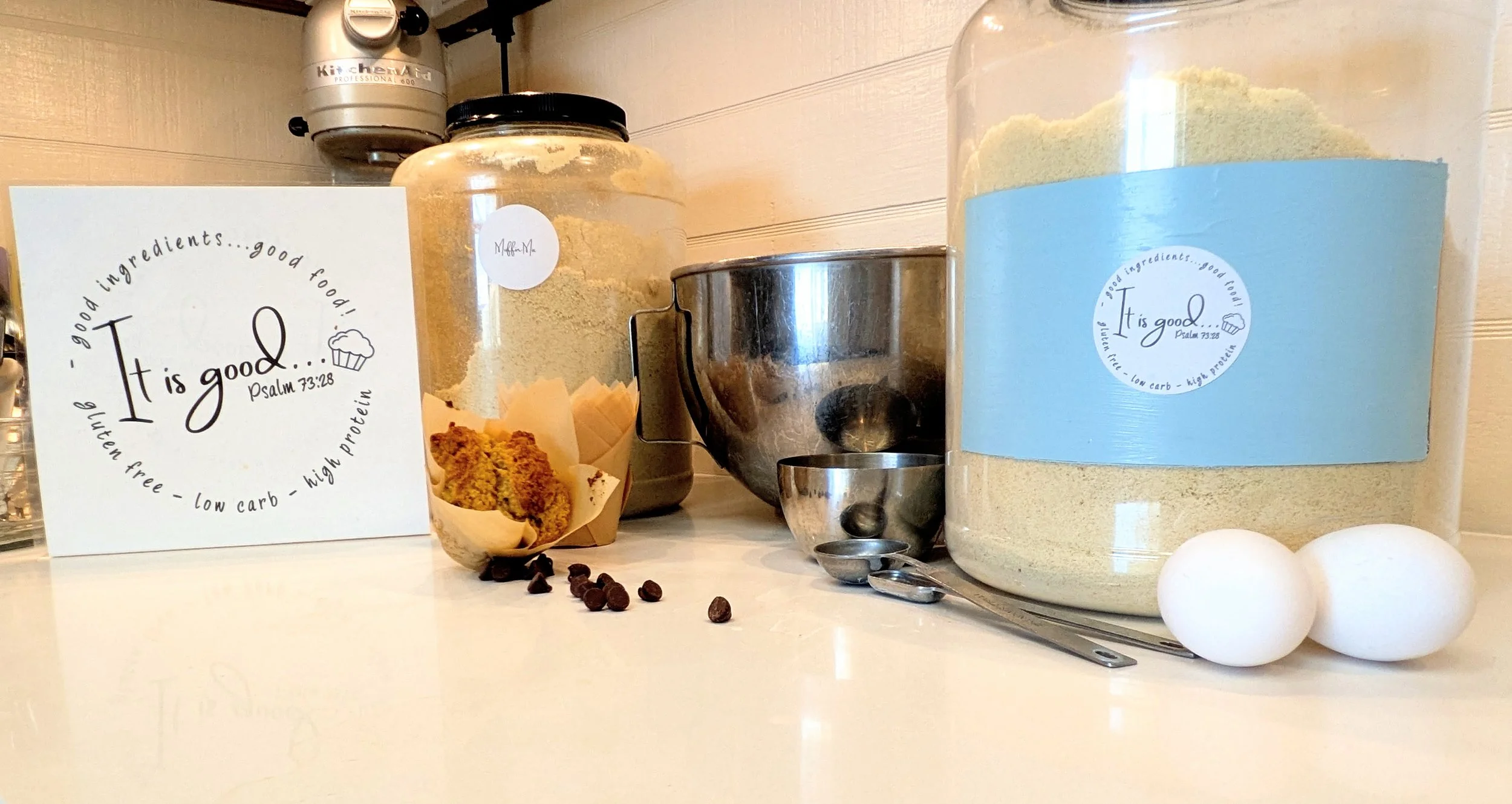 Containers of baking ingredients on a kitchen counter, including a large jar of flour with a blue label, a jar with a black lid, and a metal mixing bowl. There is a small handwritten sign with a quote from Psalm 73:28, and baking items like eggs, chocolate chips, and measuring spoons are also visible.