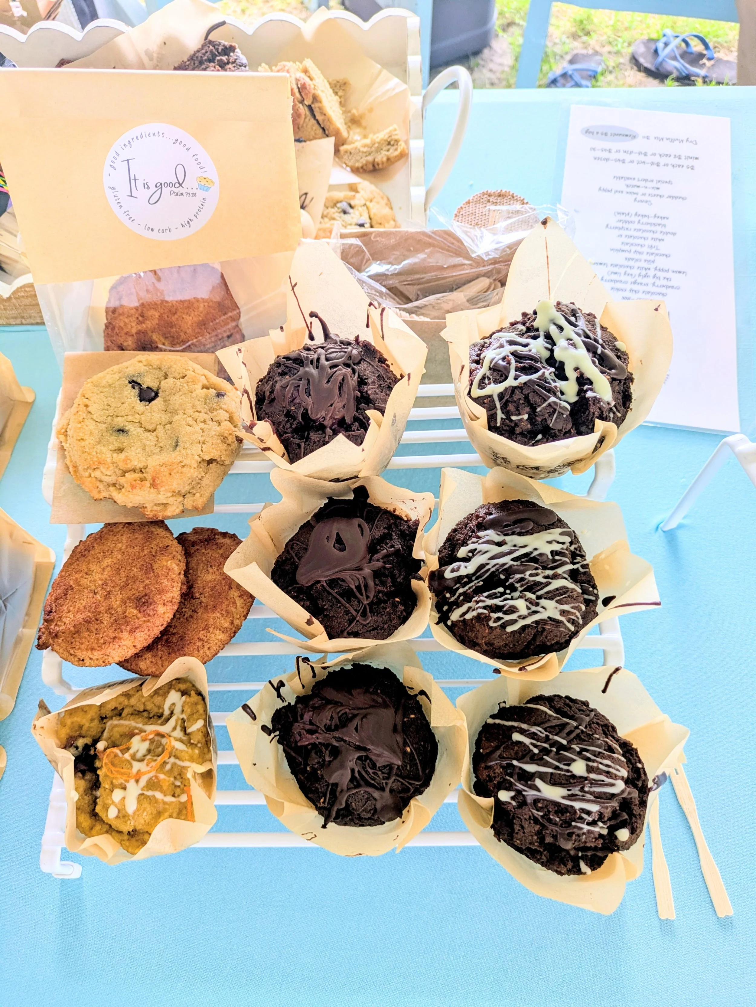 Assorted cookies and muffins on a white rack, with some wrapped in paper cups, some topped with chocolate or white drizzle, and a background of a blue table and outdoor grass.