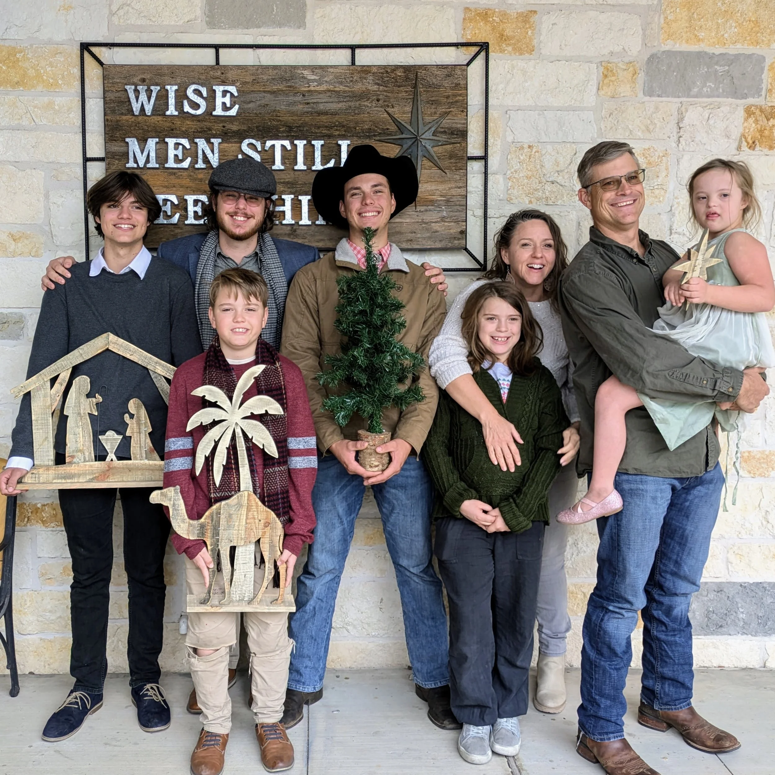 Group of people, including children and adults, standing in front of a stone wall with a wooden sign that reads 'Wise Men Still Seek Him' and holiday decorations such as a small Christmas tree and wooden cutouts.