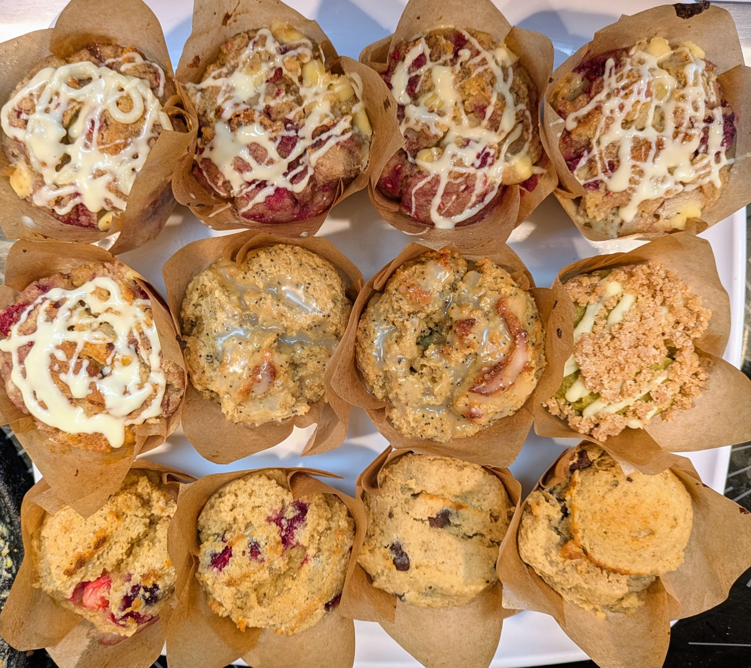 A variety of muffins in paper cups, topped with drizzles of white icing, on a white platter.