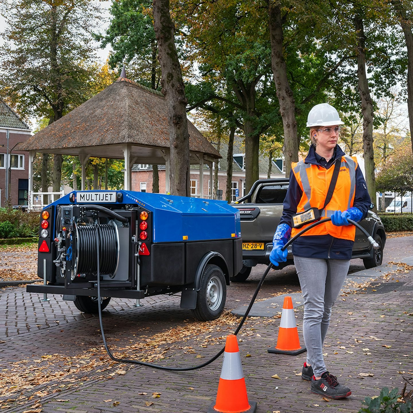 A female worker wearing safety gear and gloves operating high-pressure cleaning equipment on a paved surface in a park with trees and buildings in the background.