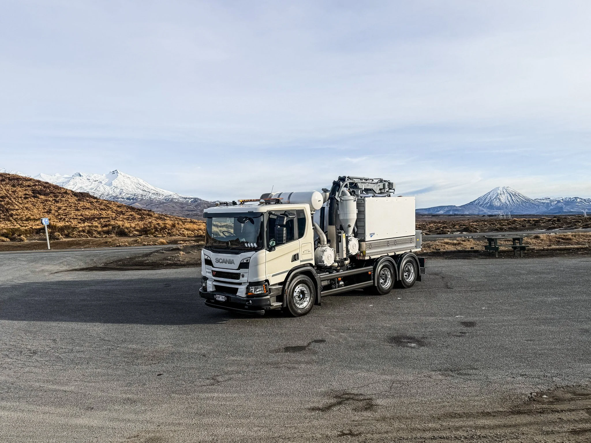A white service truck with equipment on top, parked on the side of a road with a mountainous landscape in the background, including snow-covered peaks and a partly cloudy sky.