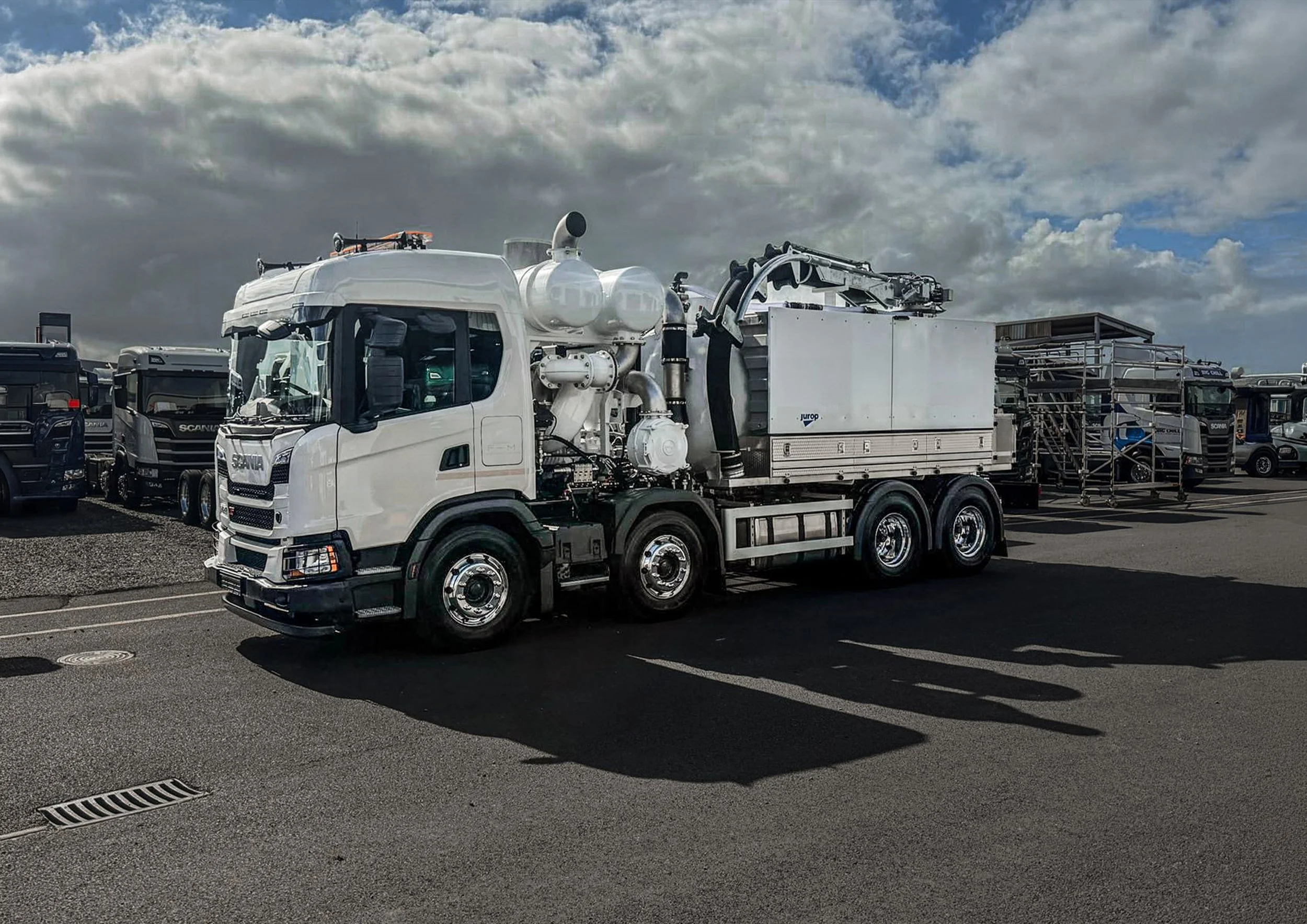A white truck equipped with machinery and equipment for cleaning or servicing, parked in a lot filled with other trucks and vehicles, under a cloudy sky.