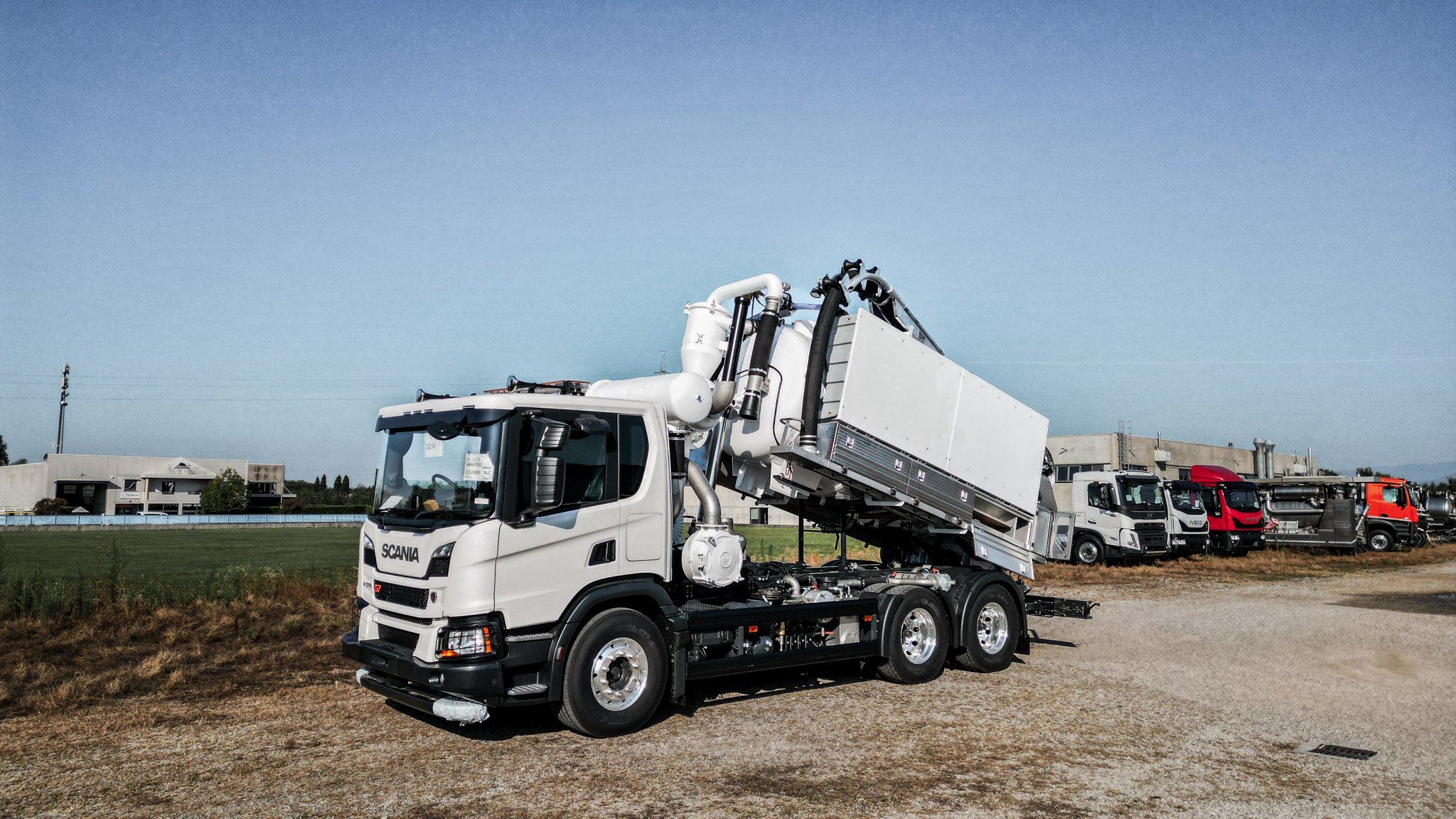 Line of trucks parked in a lot, including a white truck in the foreground with a mounted crane or arm, and buildings in the background under a clear sky.