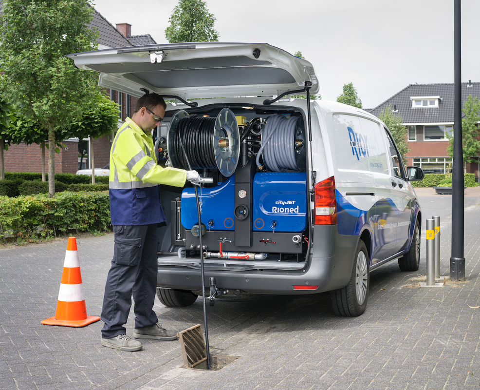 Technician in safety vest inspecting underground sewer or drain pipe connected to a utility service vehicle with hoses, on a city street.