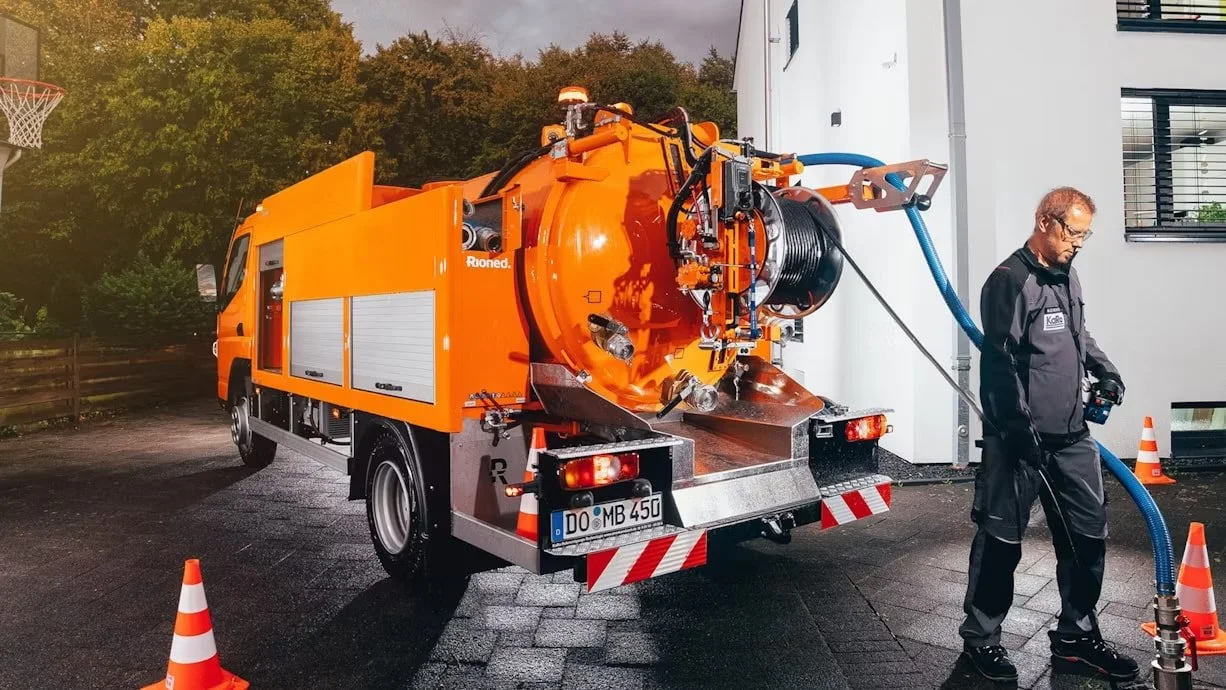 A person in black work overalls standing next to an orange vacuum truck with a large hose connected to a building, with traffic cones around for safety.
