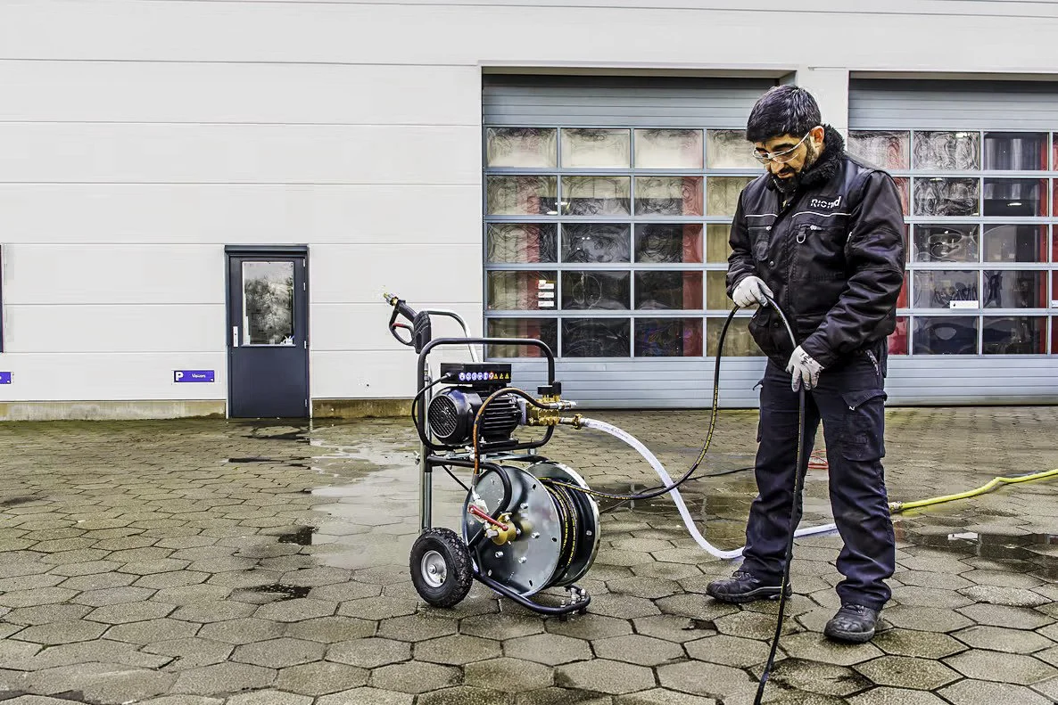 A man wearing glasses and gloves is operating a pressure washer outside a building, cleaning a paved area.