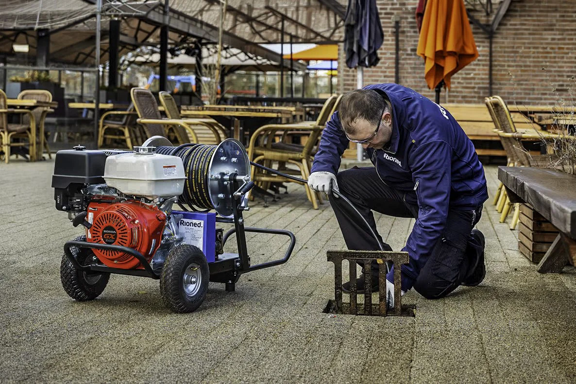 A man in a blue jacket kneels next to an open outdoor drain, working with a drain snake machine to clear a sewer or drain blockage. The machine has a red engine and a reel with a long cable. The setting appears to be a patio or outdoor seating area with tables and chairs.