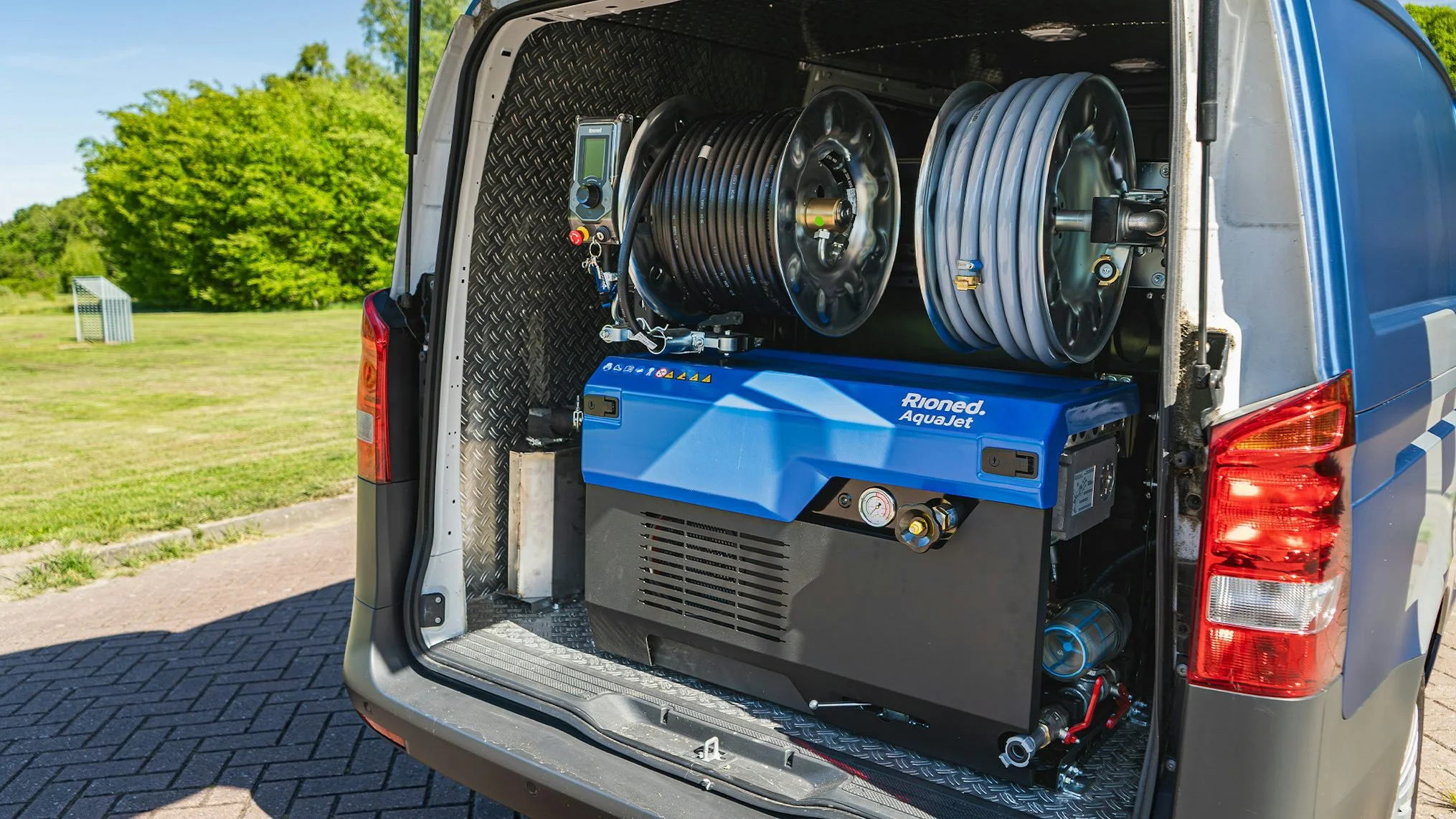 Open van door revealing equipment including hoses, a blue machine labeled 'Rioned AquaJet,' and other tools for cleaning or maintenance, set outdoors with trees and grass in the background.