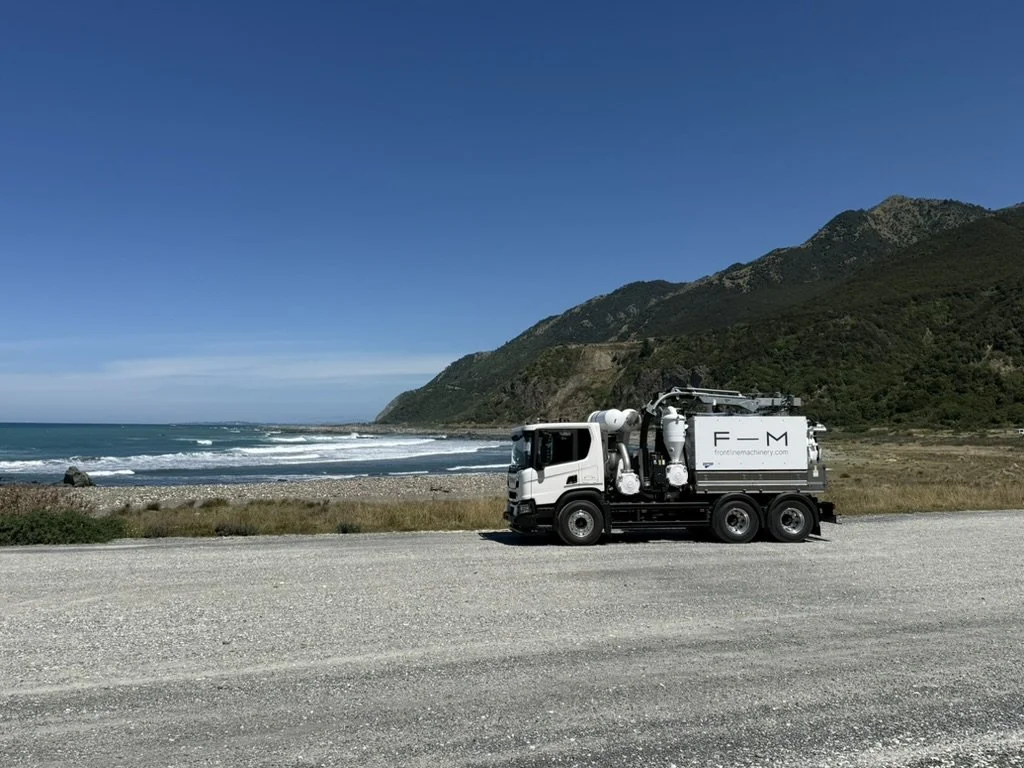 A utility truck parked on a gravel road near the coast with green hills in the background and an ocean view.