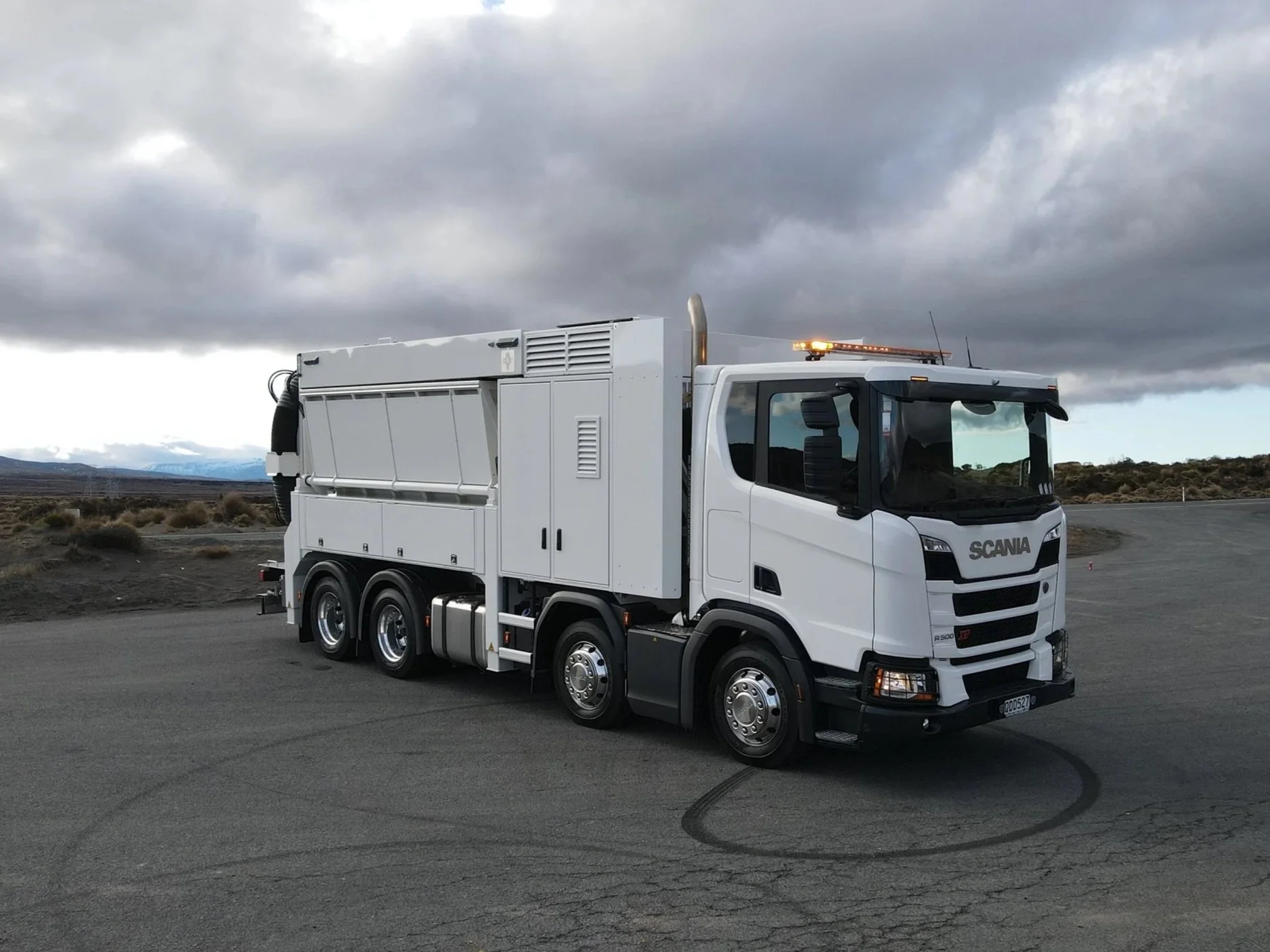 A white Scania utility truck with a specialized cargo area parked on an empty paved lot with tire marks, under a cloudy sky during daytime, with mountains and sparse vegetation in the background.
