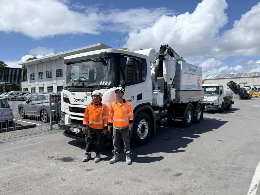 Two workers wearing orange safety vests and white helmets standing in front of a white industrial truck in a parking lot.