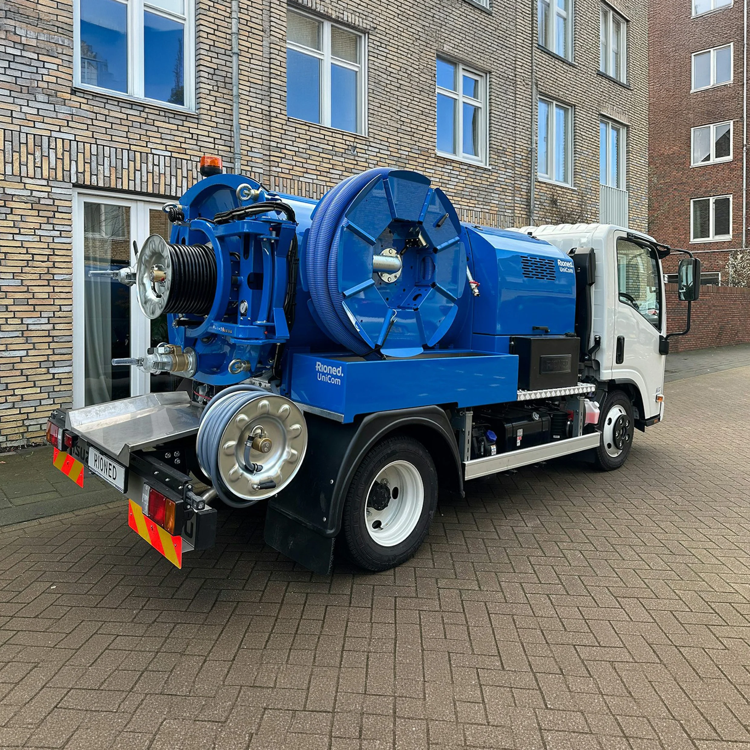 A large blue vacuum truck parked in front of a residential brick apartment building with multiple windows.