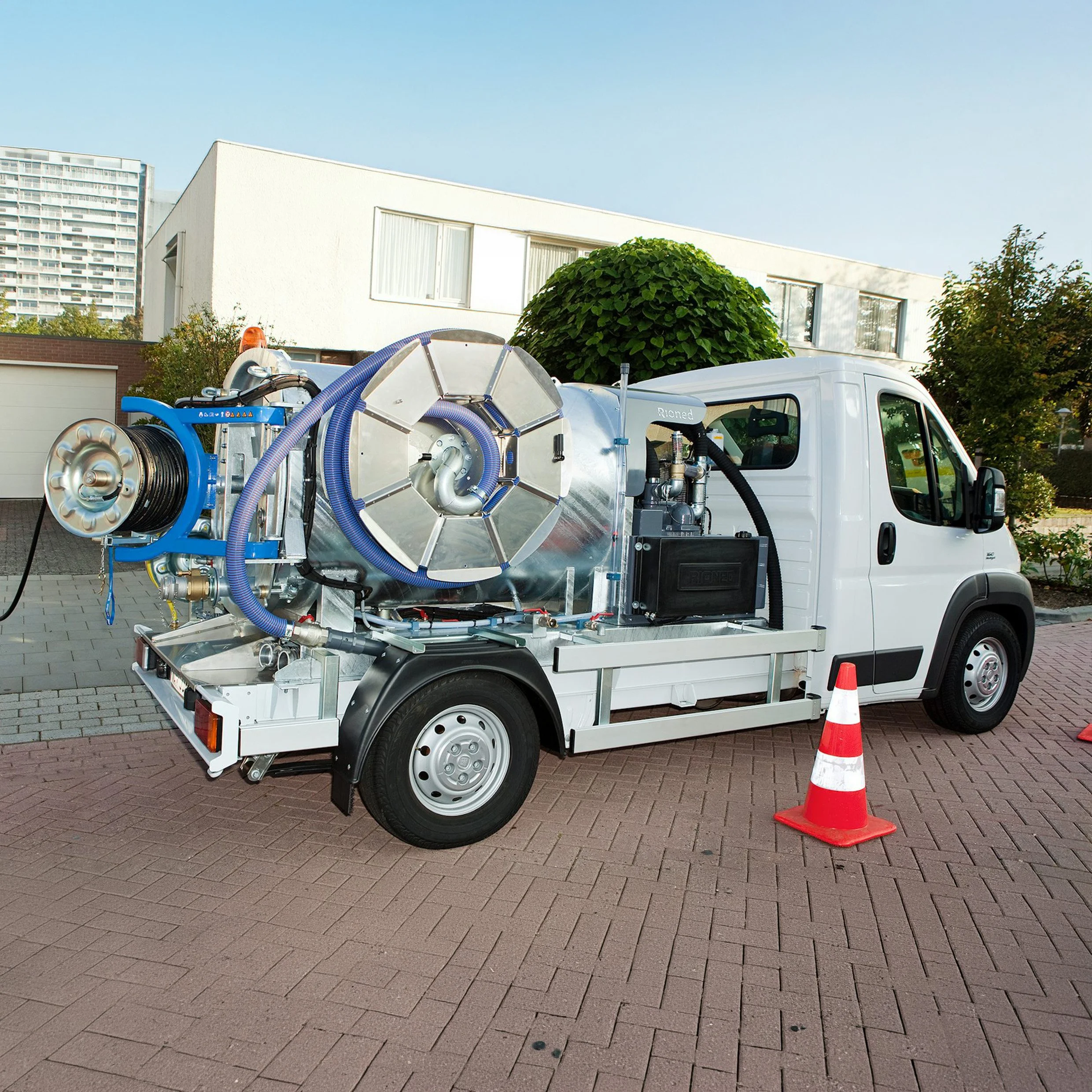 A white utility truck parked on a brick driveway with equipment and hoses mounted in the back. A traffic cone is placed near the truck, and residential buildings with trees are visible in the background.