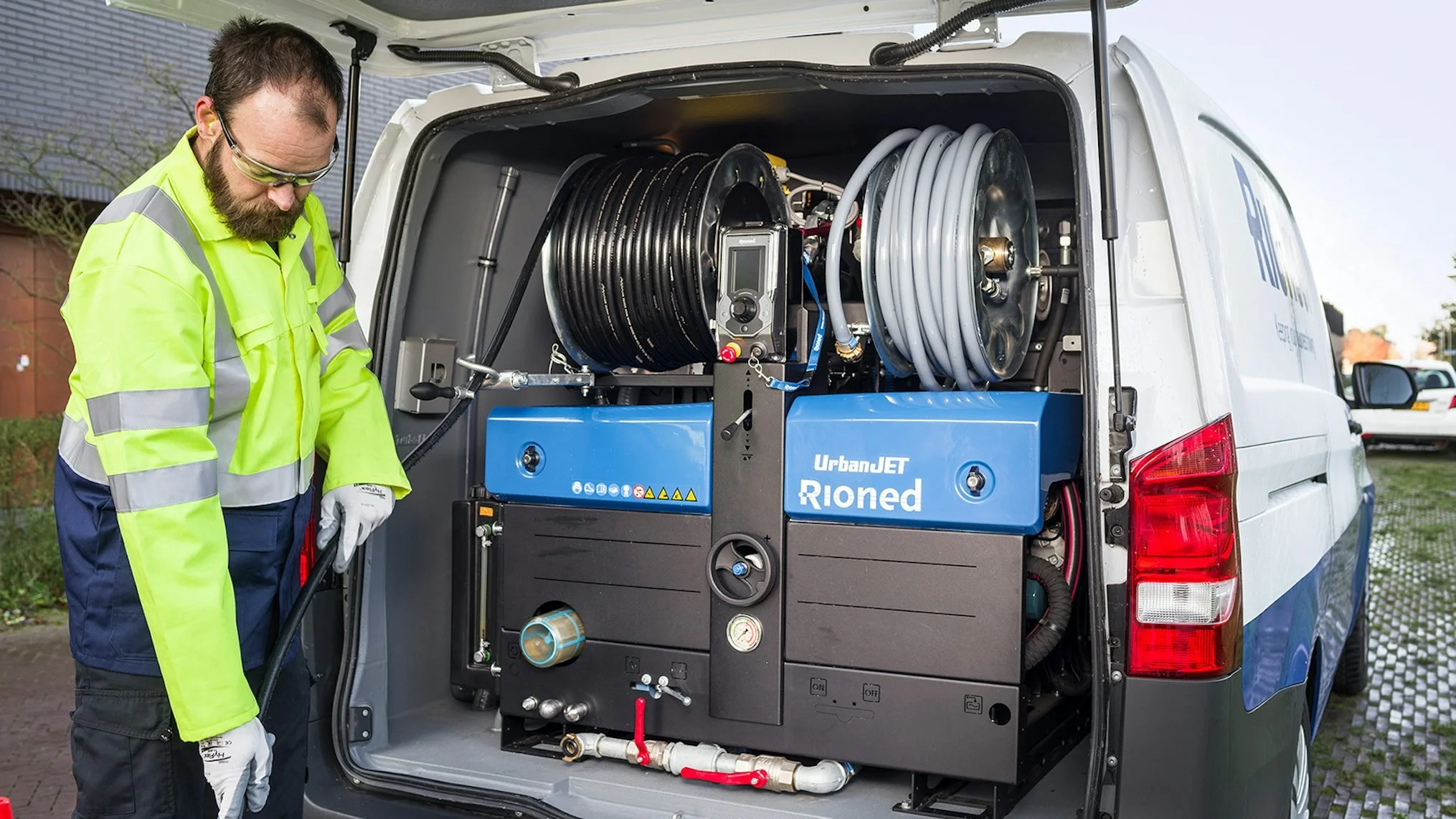 A technician wearing safety glasses and a neon yellow jacket inspecting equipment inside an open service van during daytime.