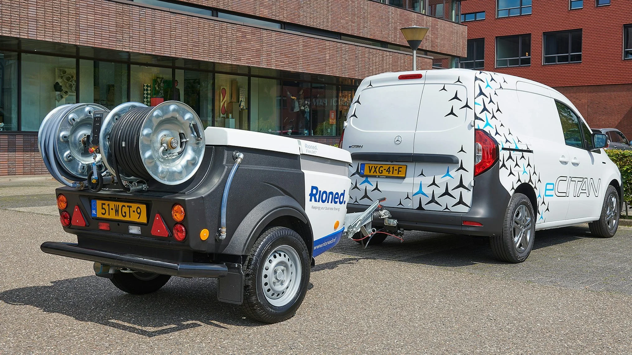 A white electric delivery van with a pattern of black and blue airplane icons on the side, attached to a small trailer carrying equipment, parked outside a modern brick building.