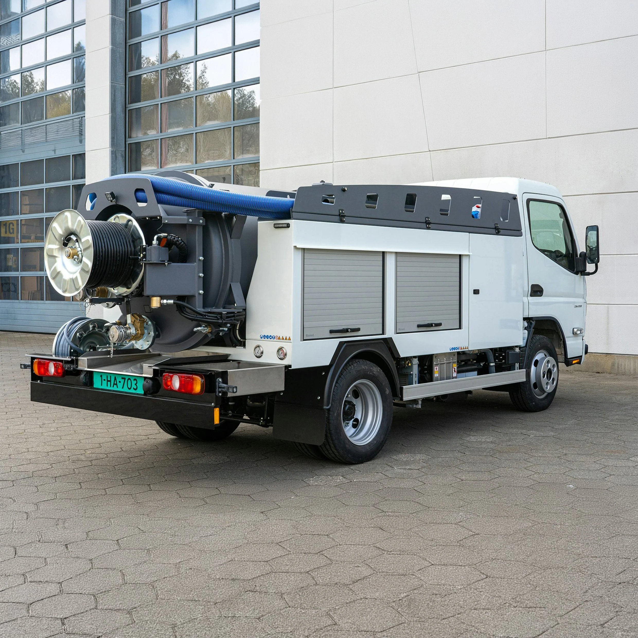 A street cleaning vehicle equipped with hoses and reels parked in front of a modern glass building with a light-colored wall.