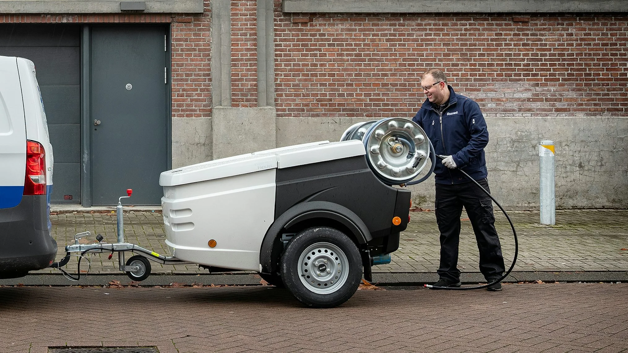 A man wearing glasses and a navy jacket is cleaning a mobile pressure washer or generator on wheels outdoors. The washer is connected to a vehicle with a trailer hitch, and he is holding a hose. The background features a brick wall, a dark grey door, and a concrete post with a yellow tape or marker. The ground is paved with bricks, and a small red-topped pole is visible in the scene.