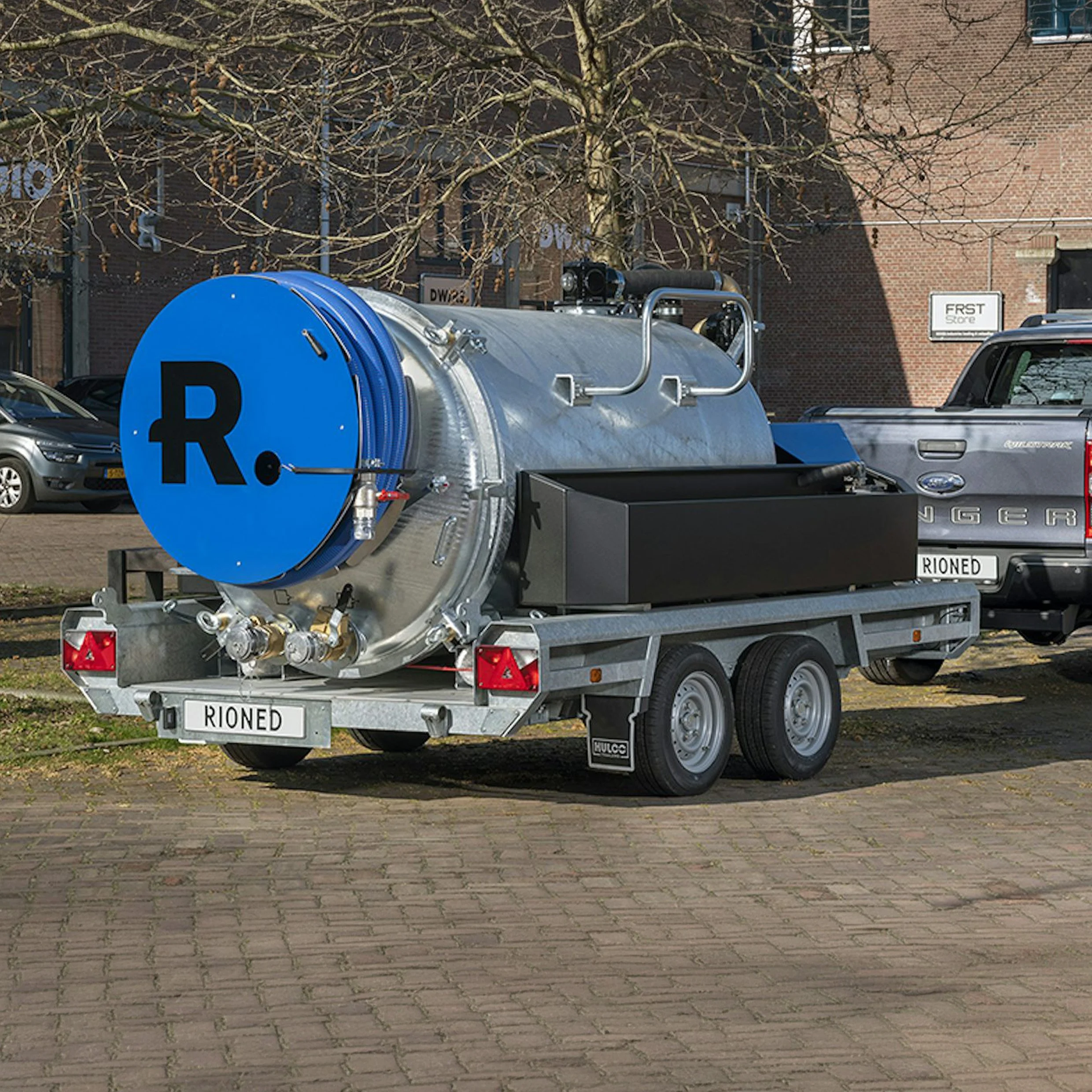 A street scene with a large silver water tank mounted on a trailer, a building with brick walls and windows, a blue parking sign with a black letter R, cars parked nearby, a tree without leaves, and a sign reading 'FIRST SCORE' in the background.
