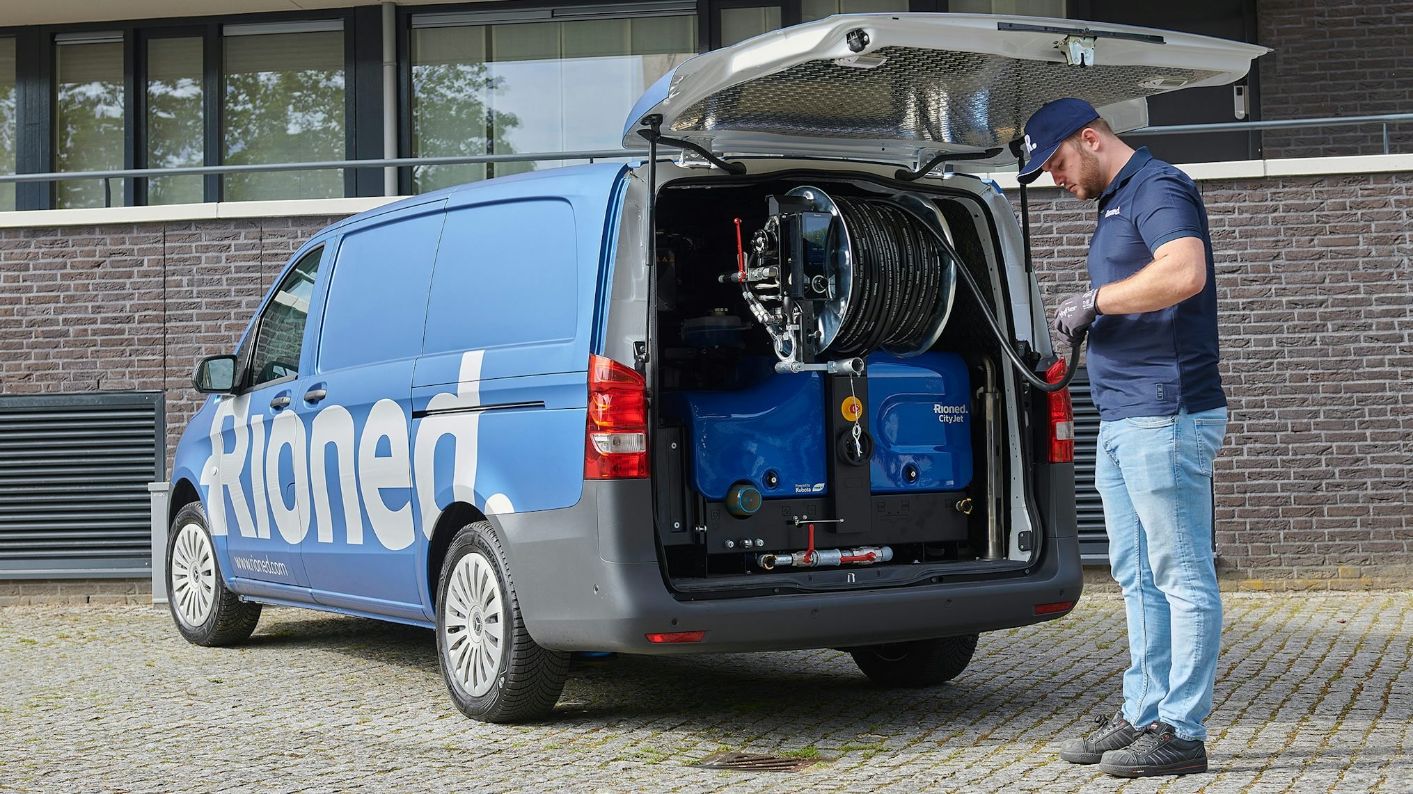 A technician in a blue uniform and cap working on a branded blue service van with its rear doors open, revealing equipment and hoses inside, in an outdoor urban setting.