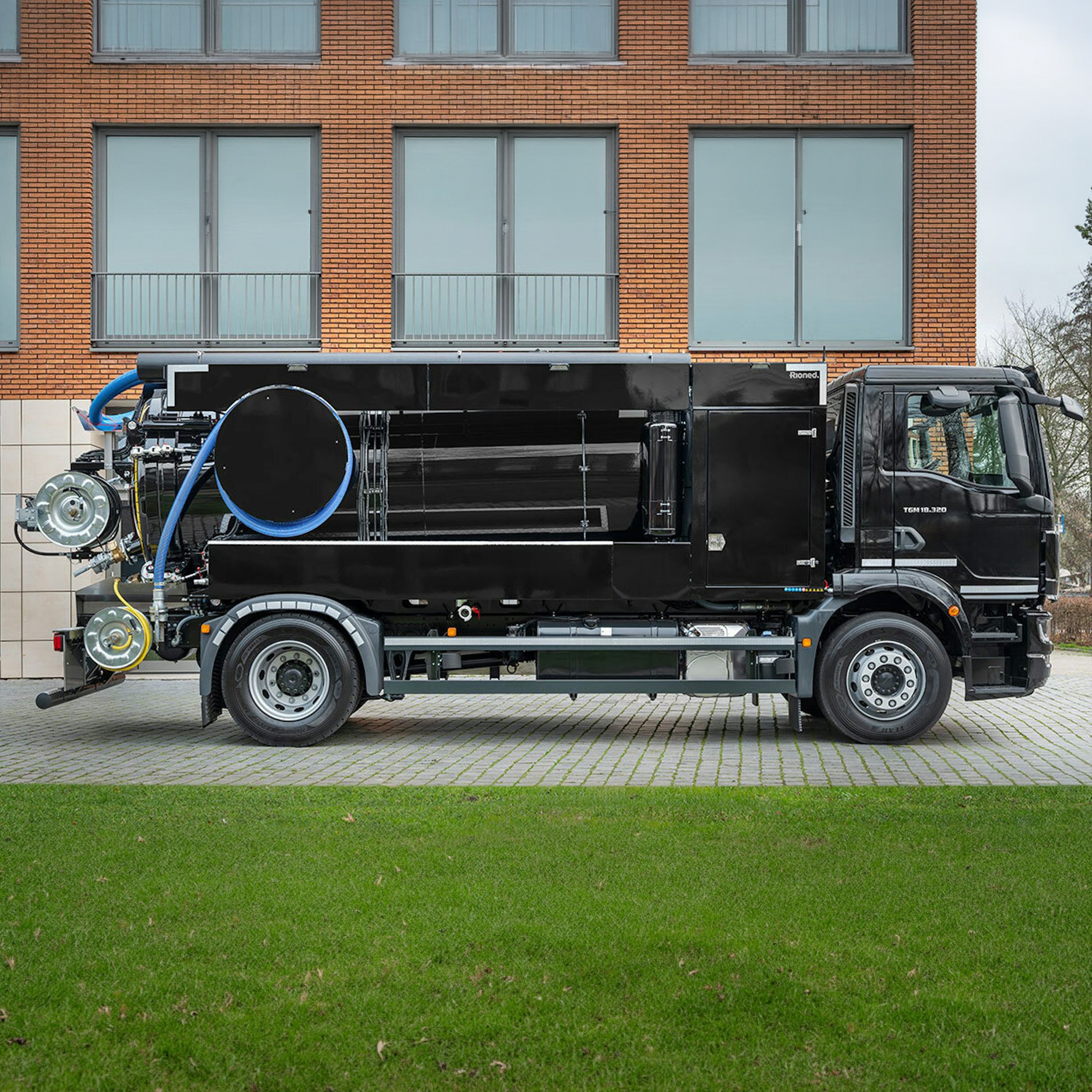 A large black truck with a flatbed trailer is parked on a paved area in front of a brick building with large windows and balconies. The truck has various equipment and hoses on its flatbed.