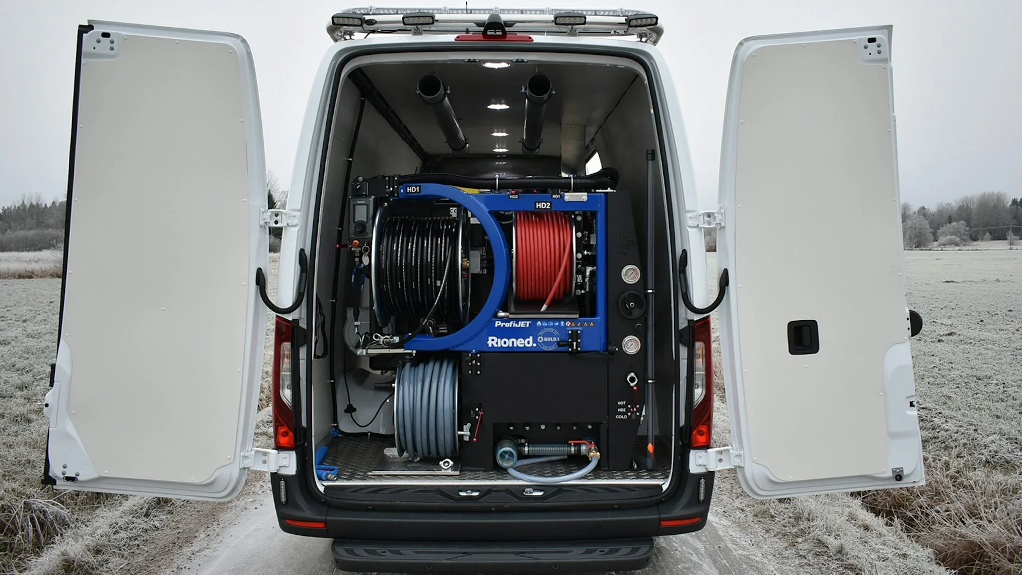 The back of a white work van with open doors revealing equipment inside, including hoses and machinery, set in a frosty outdoor area.
