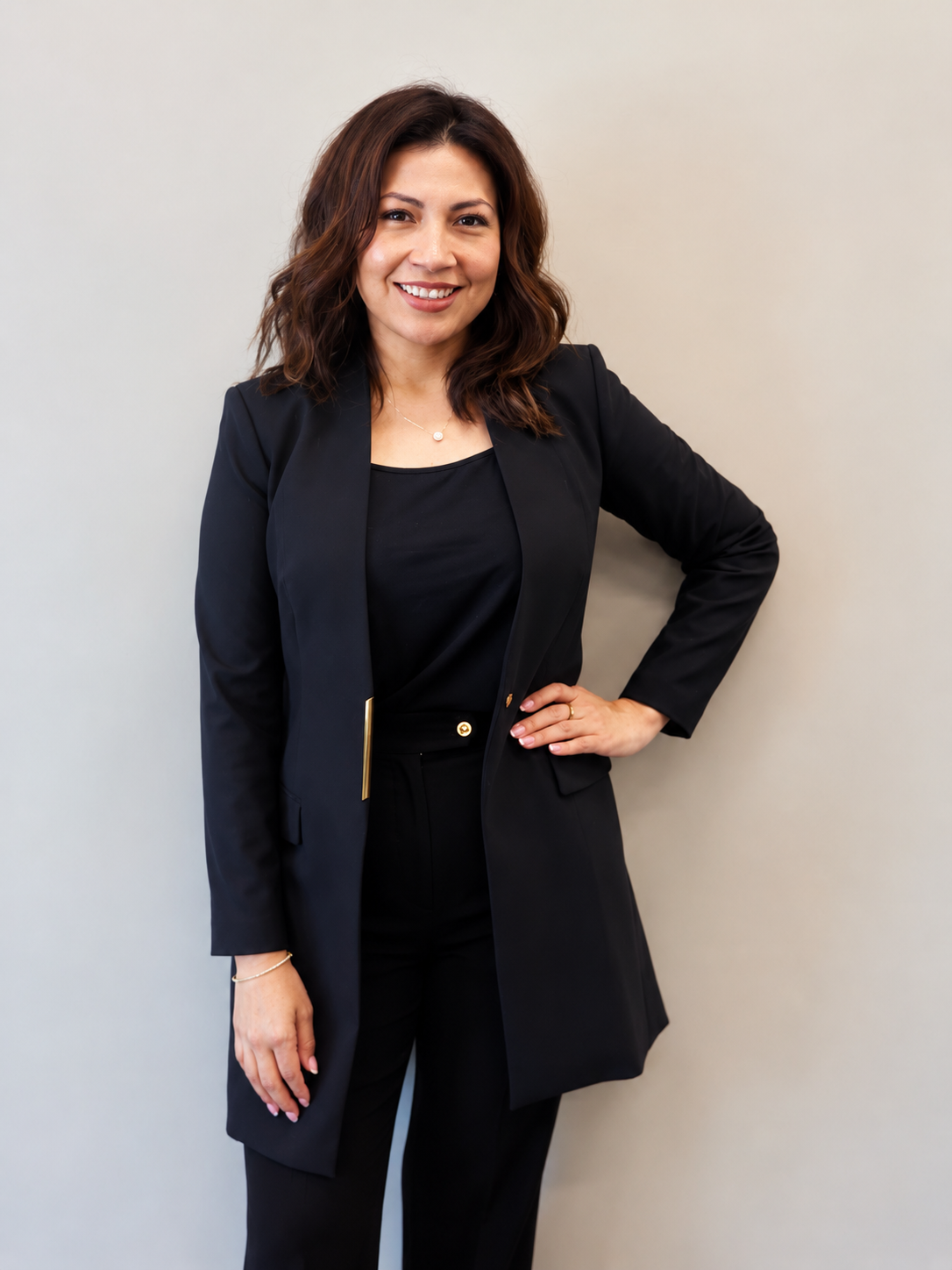 A woman with shoulder-length brown hair in a black blazer and black pants, standing against a light-colored wall, smiling, with her left hand on her hip.