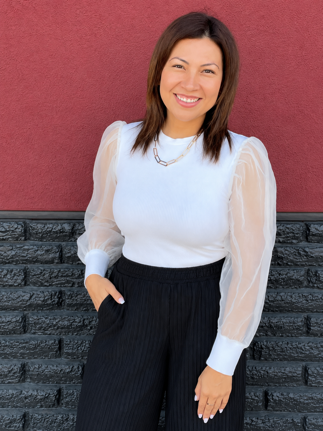 A woman smiling and standing outdoors against a red and black textured wall, wearing a white top with sheer puff sleeves, black pants, and a gold necklace.
