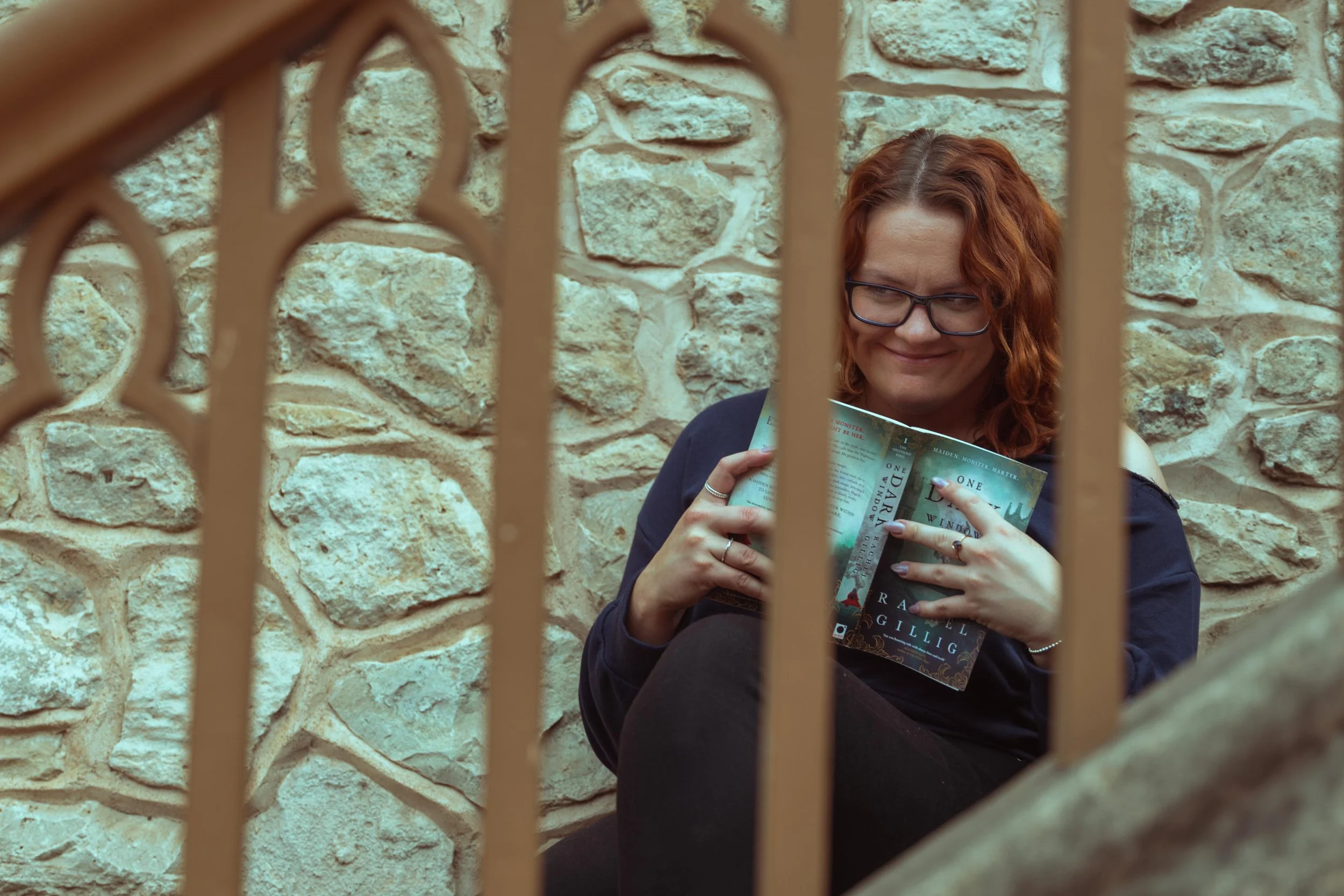Red-haired woman wearing glasses reads a fantasy novel behind iron bars against a stone wall.