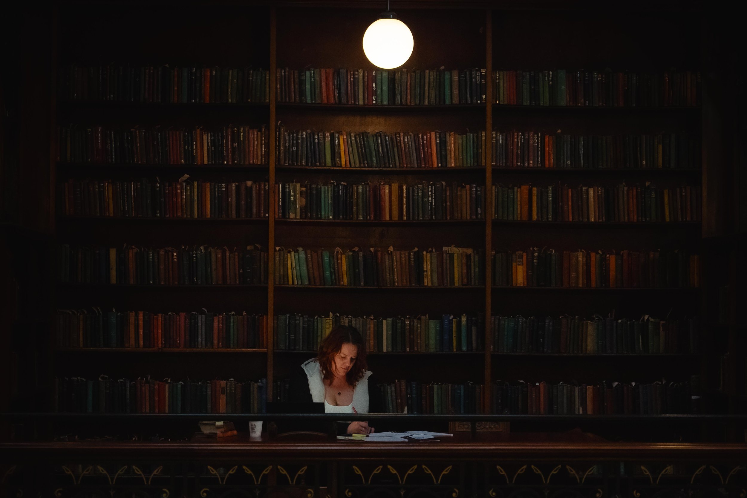 A woman with curly hair sitting at a desk with books and papers, underneath a wall of bookshelves in a dimly lit room, illuminated by a round ceiling light.