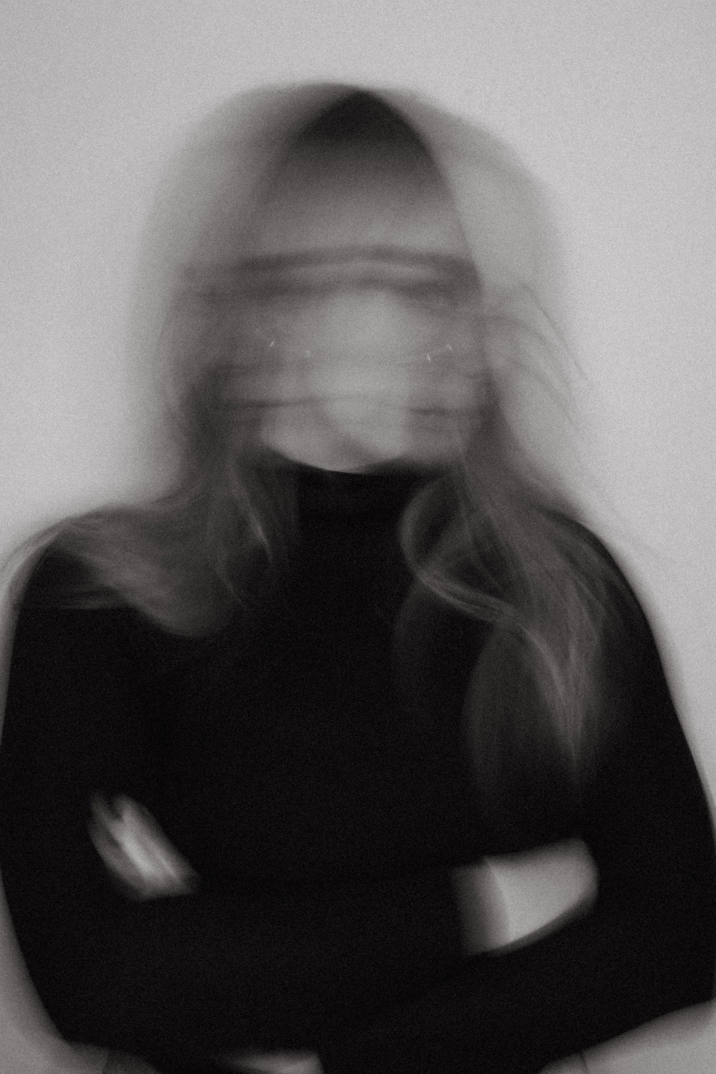 A blurry black and white photo of a woman with glasses and long hair, looking at the camera.