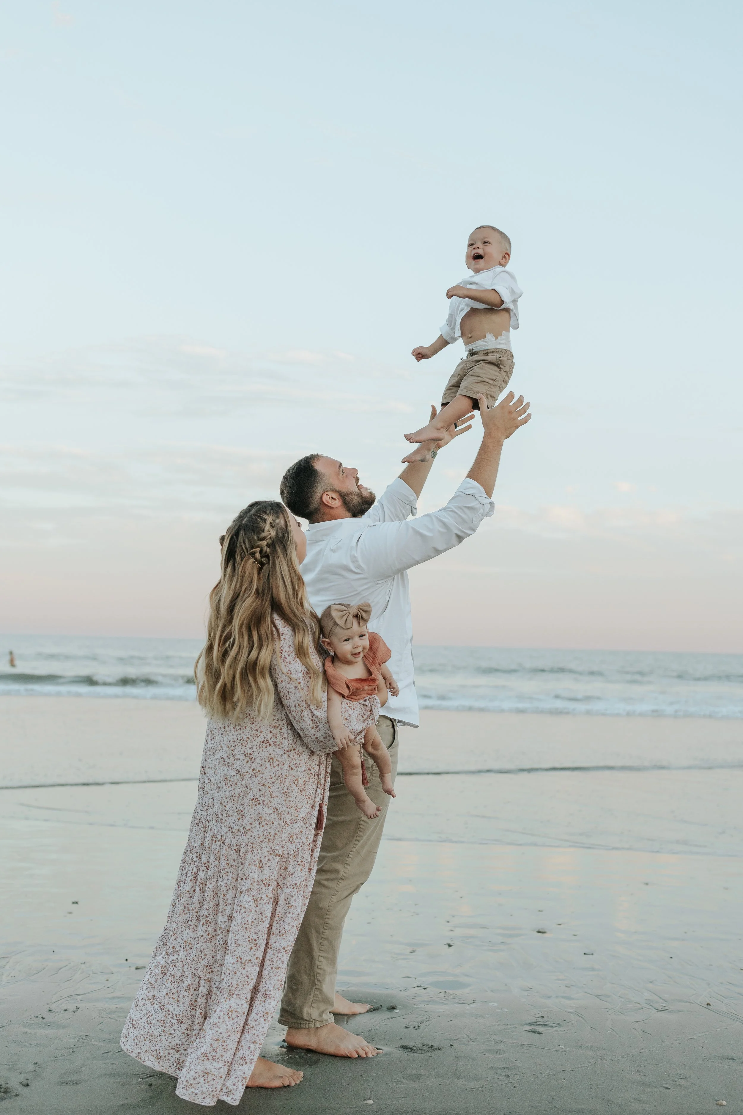 Family playing on a beach as a father lifts a laughing toddler, while a mother holds their baby.