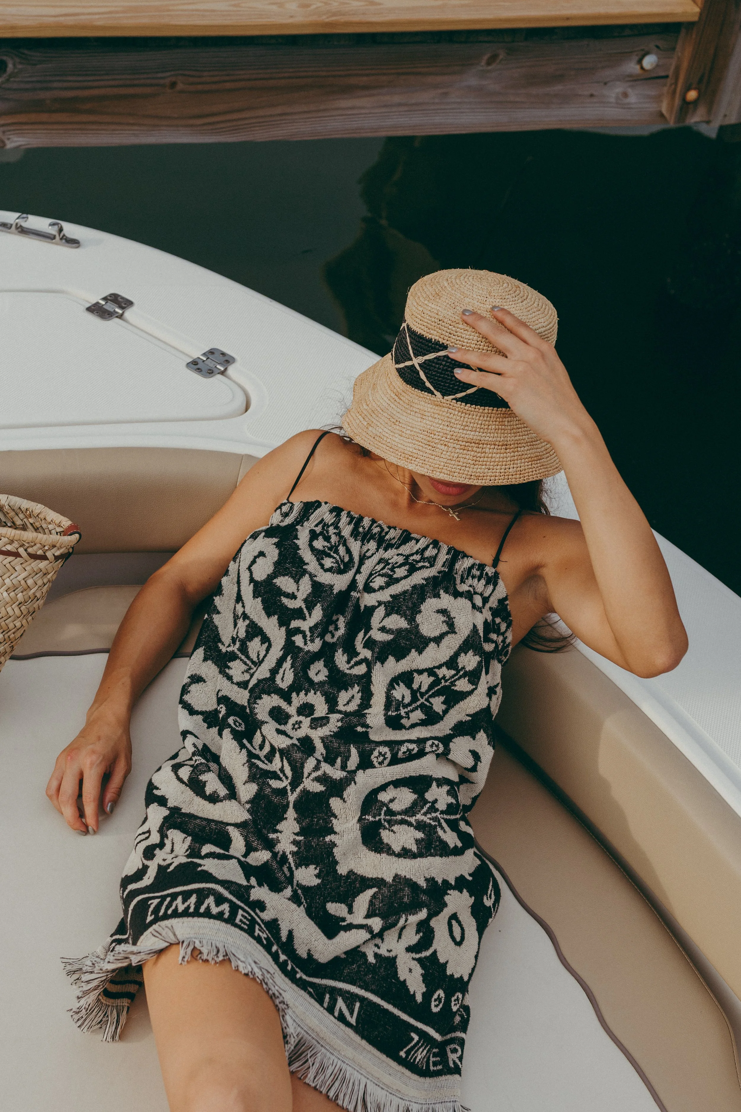 A woman in a patterned black and white towel lying on a boat with her head covered by a straw hat, her hand touching the hat, and a woven basket on the boat beside her.