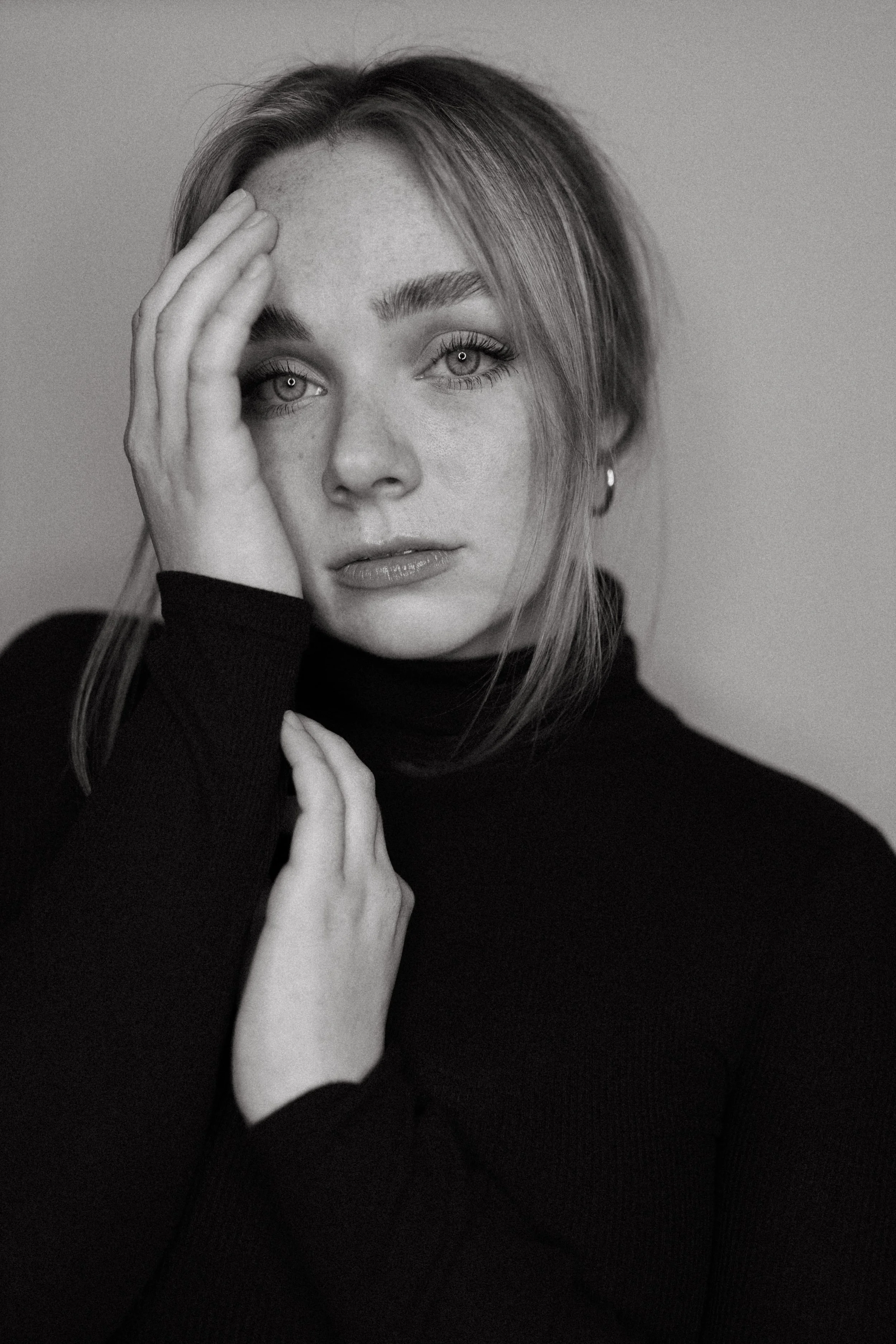 Black and white close-up portrait of a woman with light-colored eyes, holding her hand to her face, wearing a dark top and a small earring.