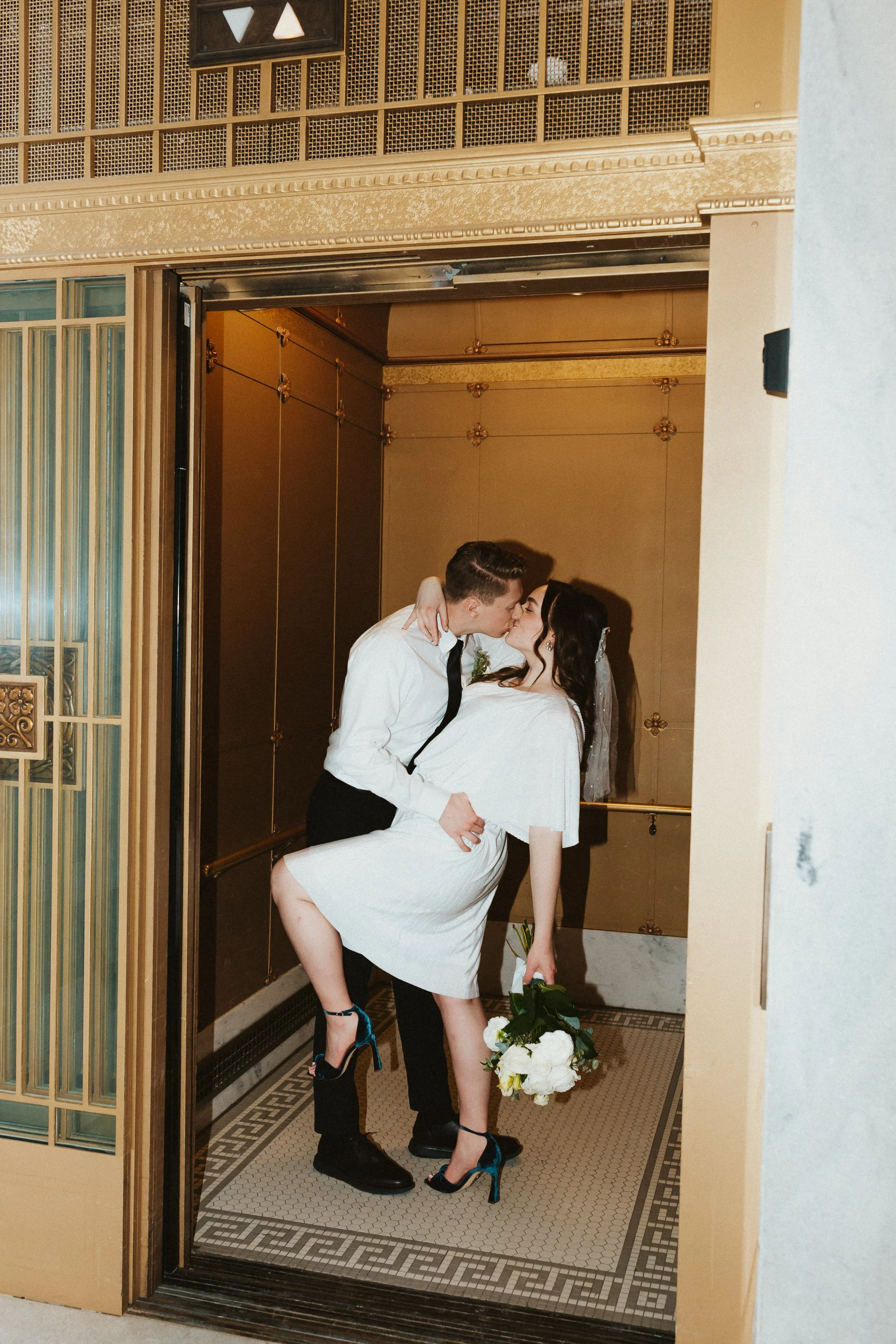 A couple sharing a kiss in an elevator with woman in white dress, holding a bouquet of white flowers.