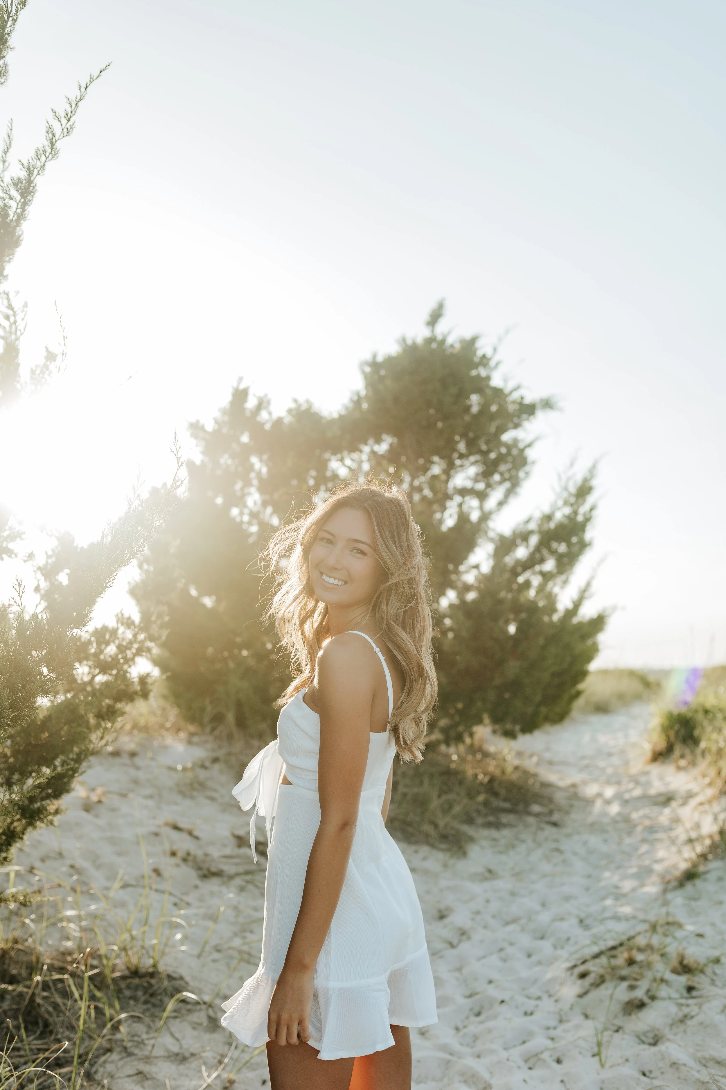 Smiling woman in a white dress standing on a sunlit sandy beach surrounded by trees