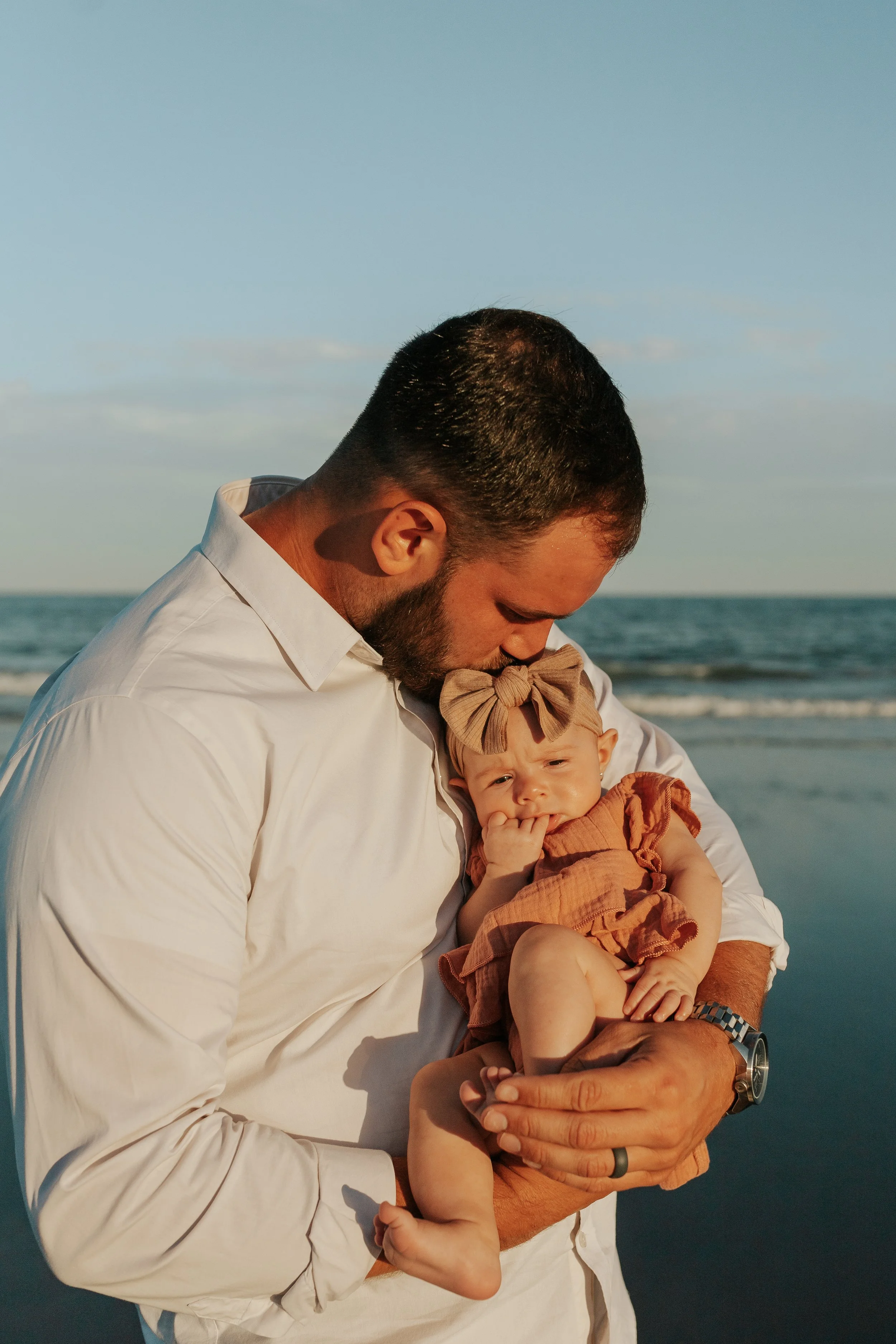 Father in a white shirt holding a baby girl in a pink dress and bow outdoors by the water