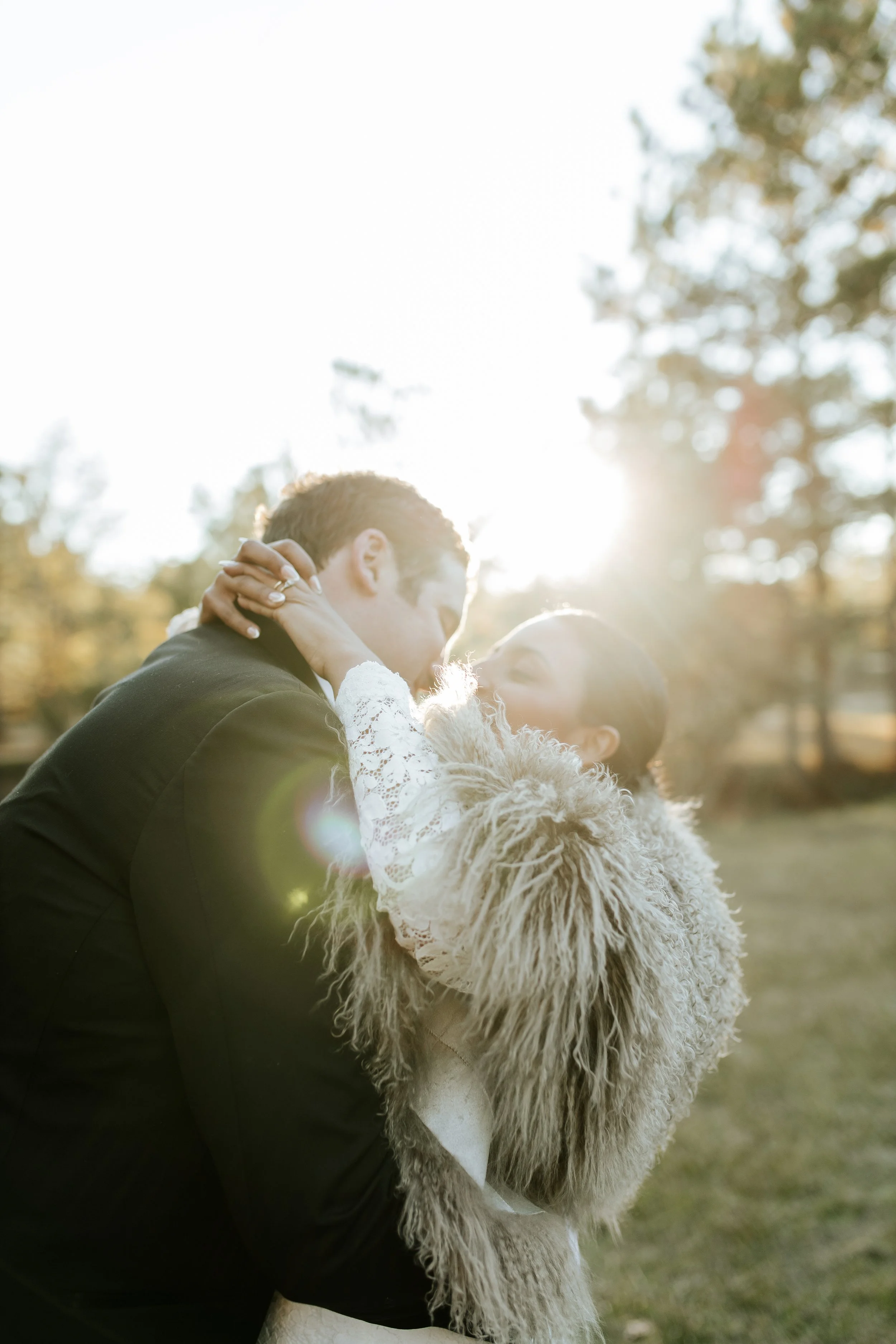 A couple sharing a kiss outdoors in the sunlight, trees in the background.