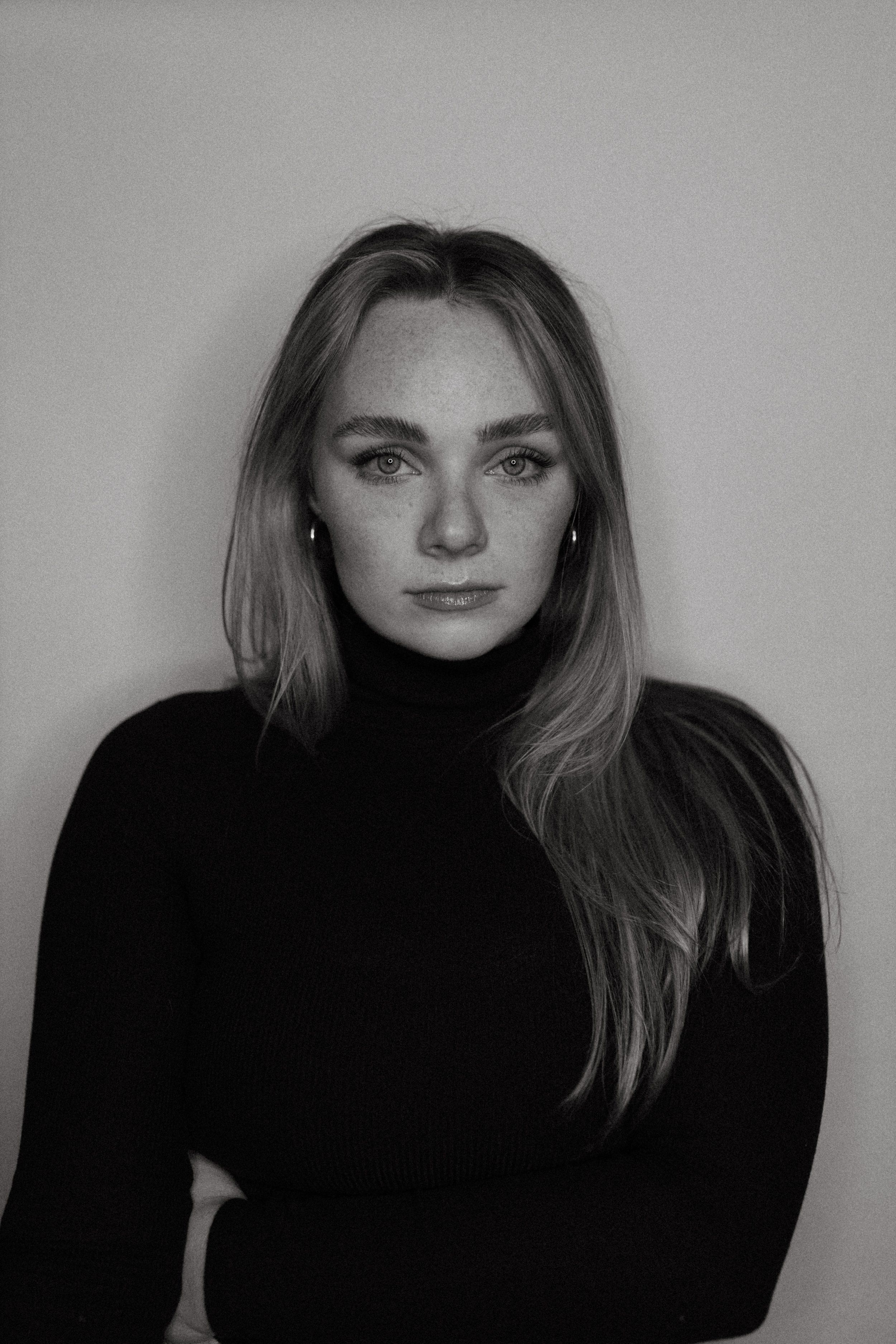 Black and white portrait of a woman with light hair, wearing a black turtleneck, looking directly at the camera, with a plain light background.