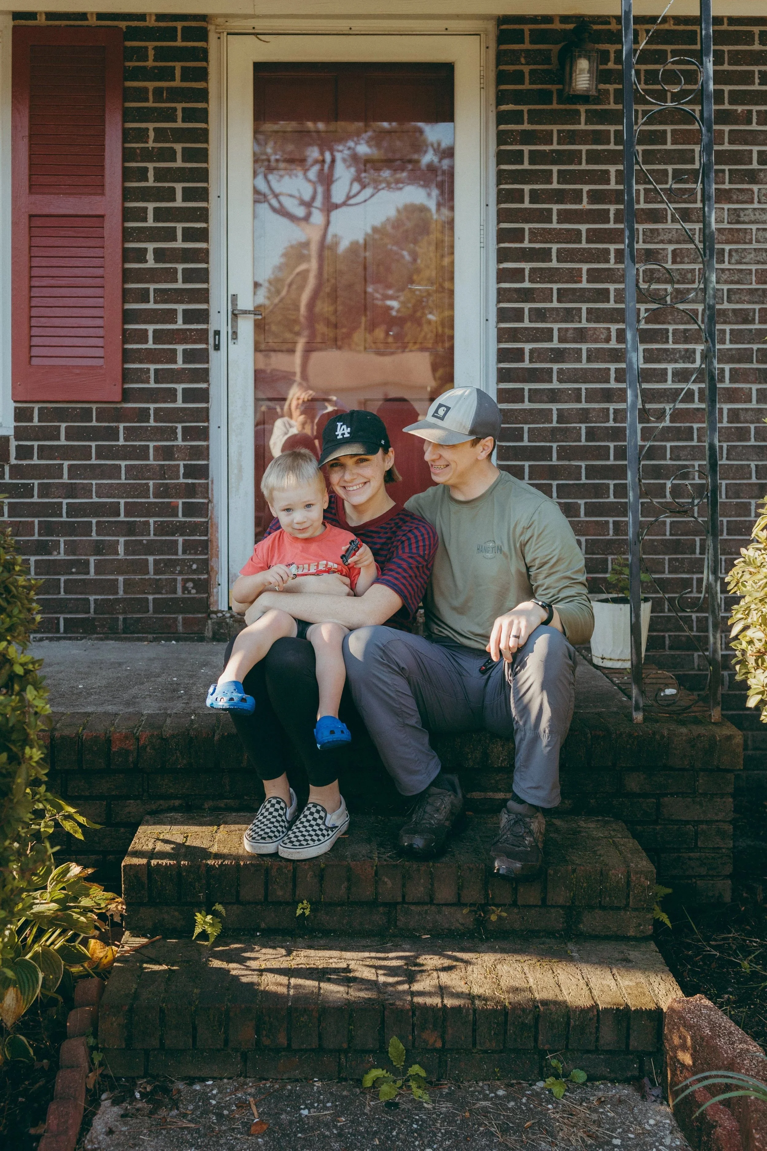 Family portrait on doorstep of brick house with couple in casual clothes and baseball caps.