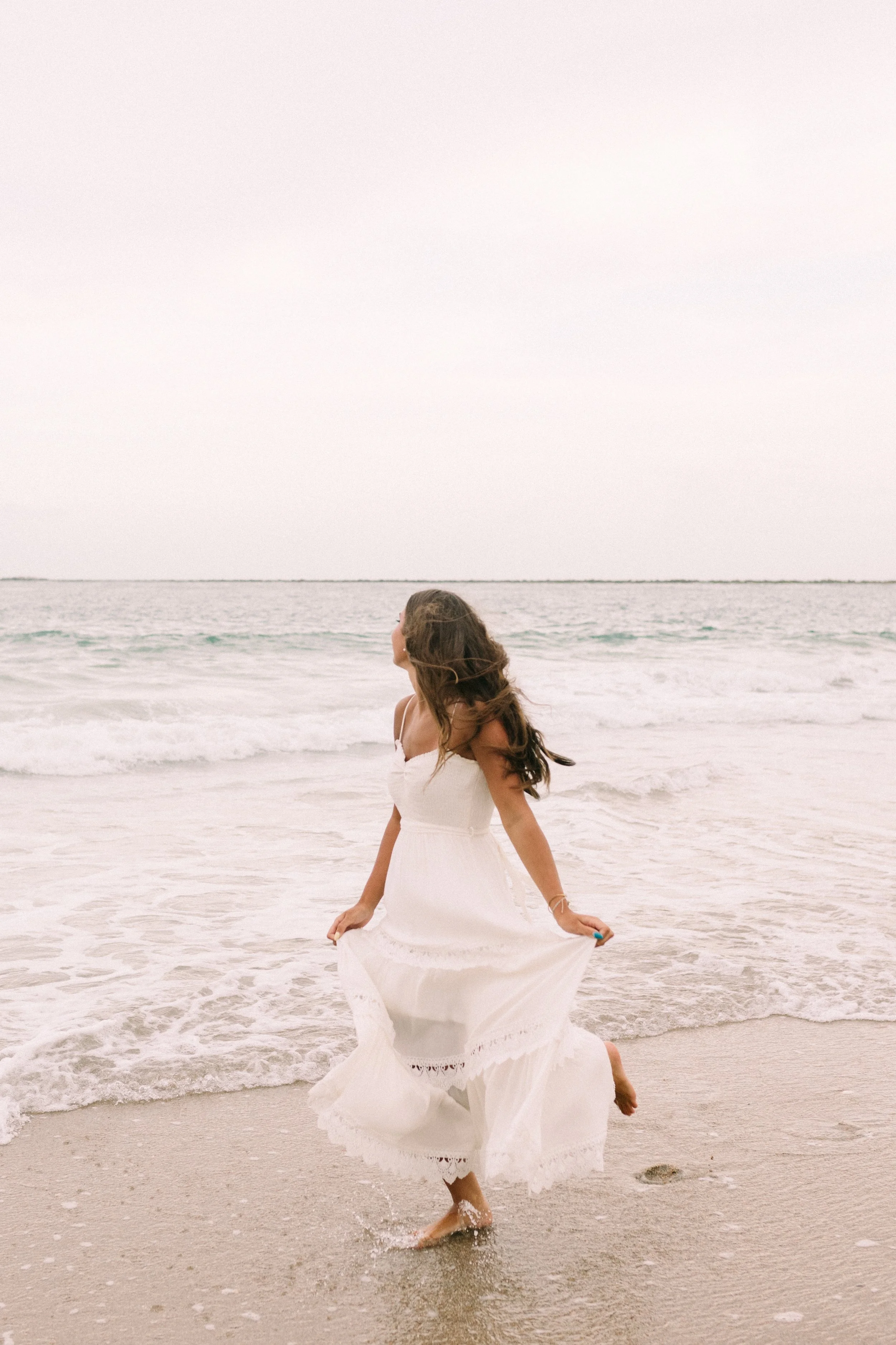 Woman in a flowing white dress walking barefoot through shallow ocean waves on a beach