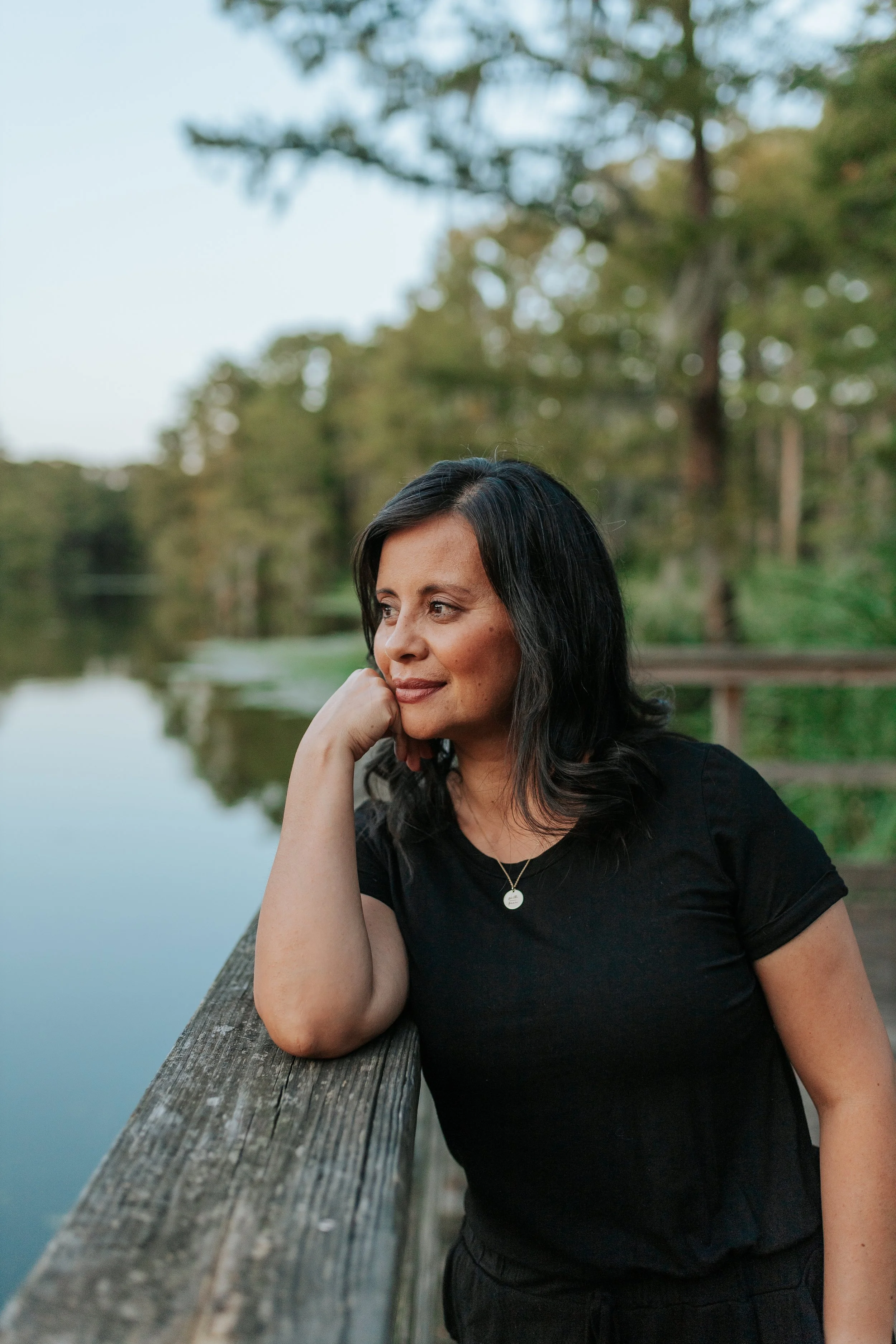 Woman in a black shirt leaning on a wooden railing beside a calm lake, with trees in the background