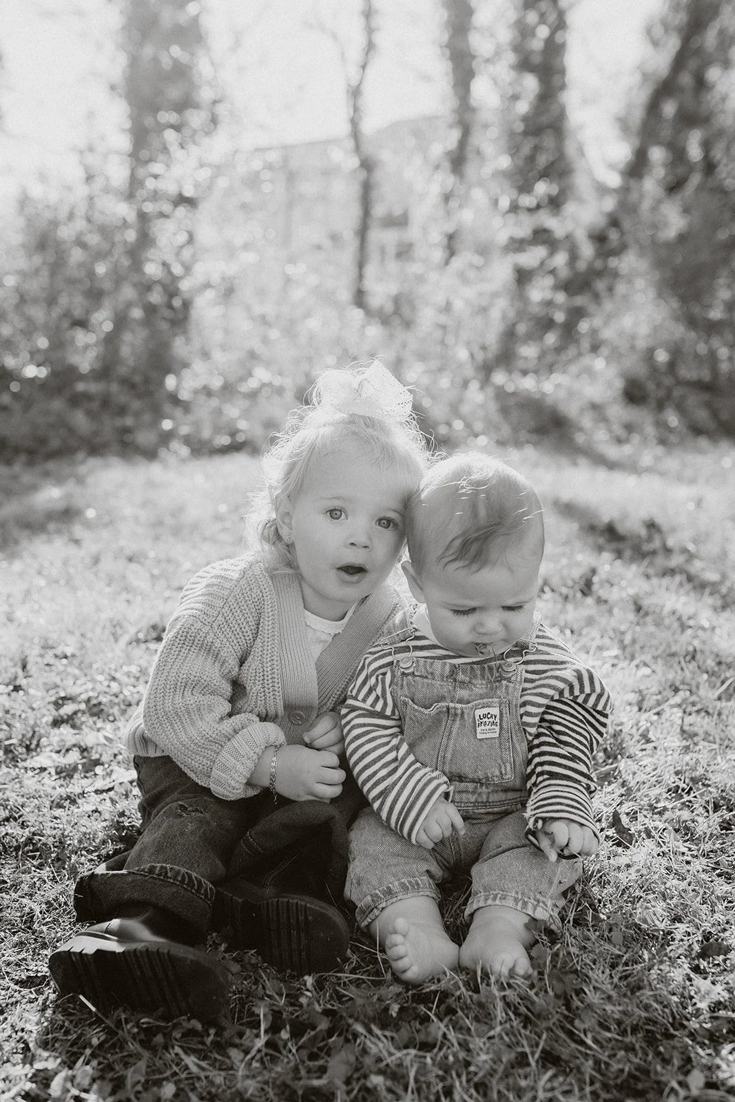 Black-and-white outdoor portrait of two young siblings sitting together on grass in a sunlit park