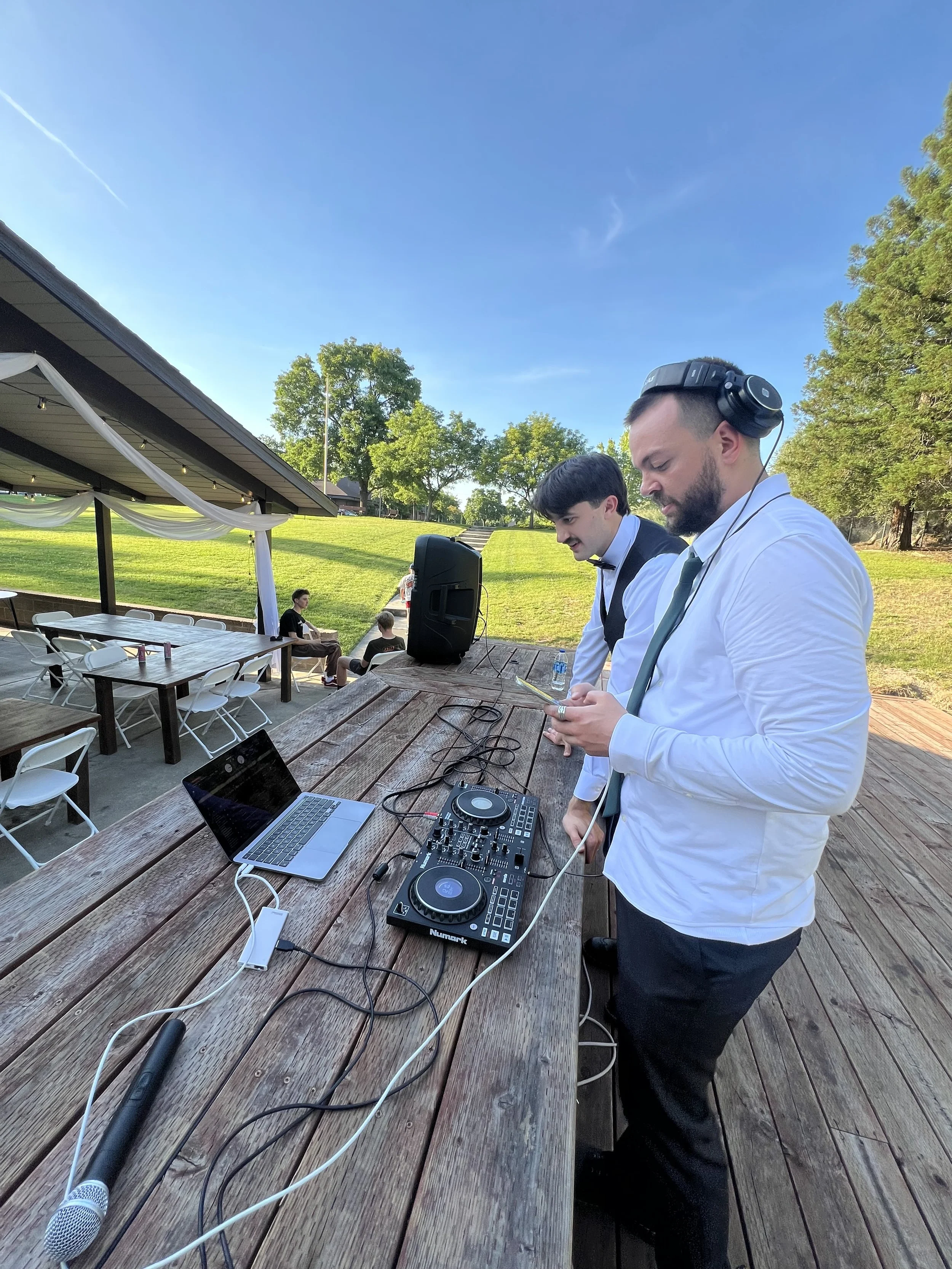 Two men in formal attire, one with headphones, DJ equipment, and a laptop on a wooden table outdoors during daytime. People seated in the background, green trees, blue sky with clouds.