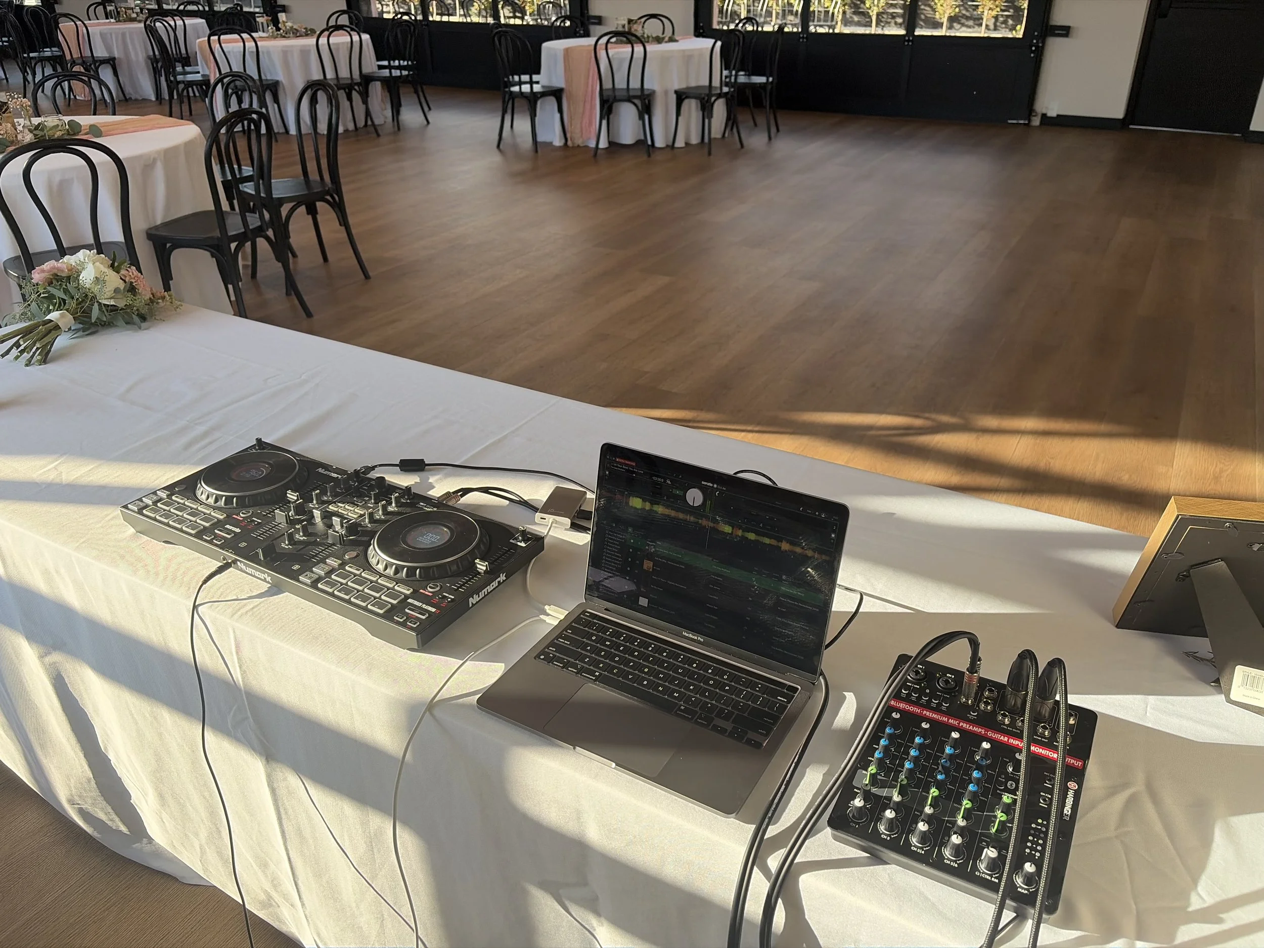 Event venue with round tables covered in white tablecloths, decorated with floral centerpieces, set up for a celebration. In the foreground, DJ equipment, including turntables, a laptop, and a mixer, is placed on a table.