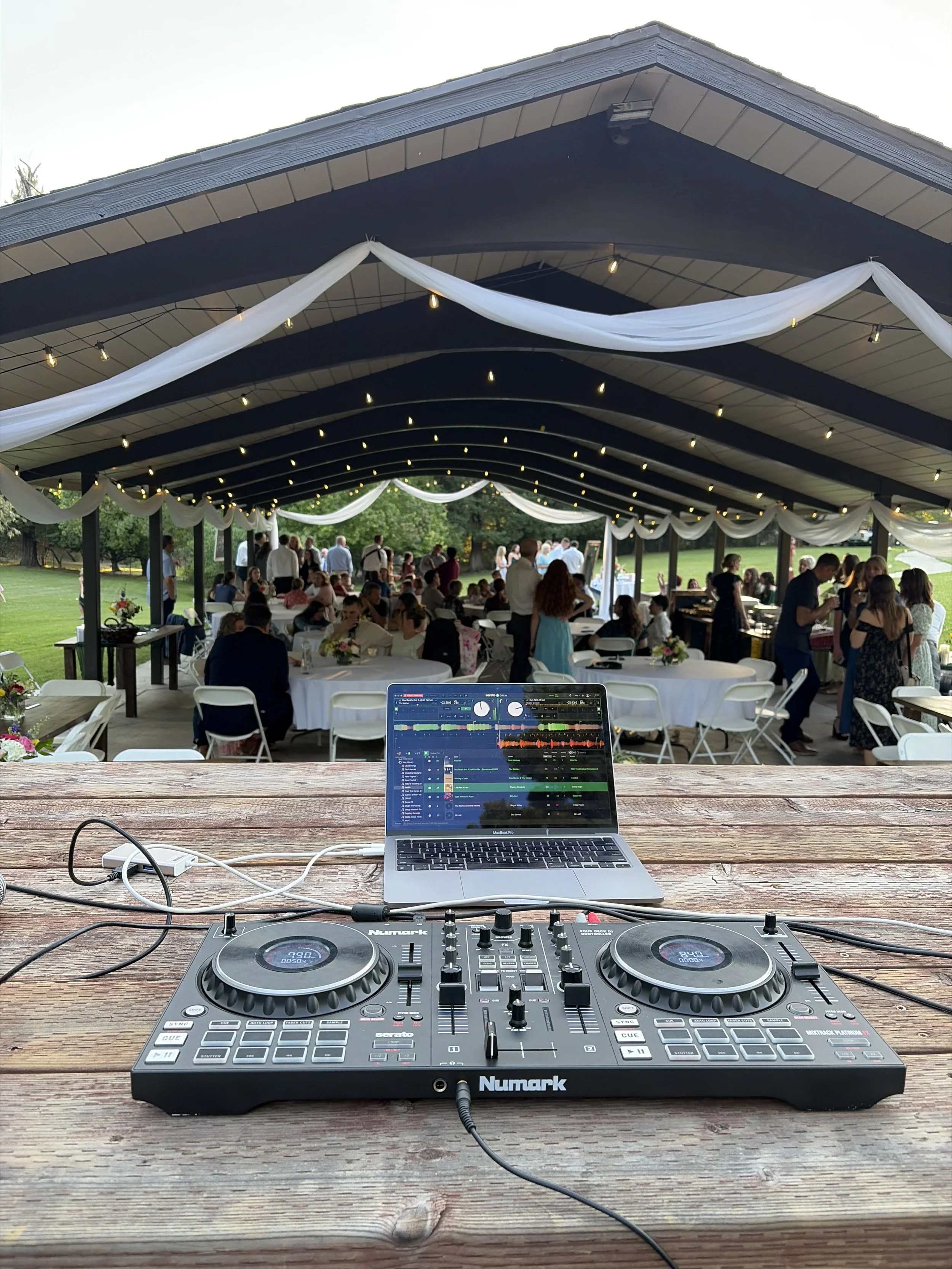 DJ equipment on a wooden table with a laptop and a large covered outdoor event space in the background showing people mingling, decorated with white drapes and string lights on a sunny day.