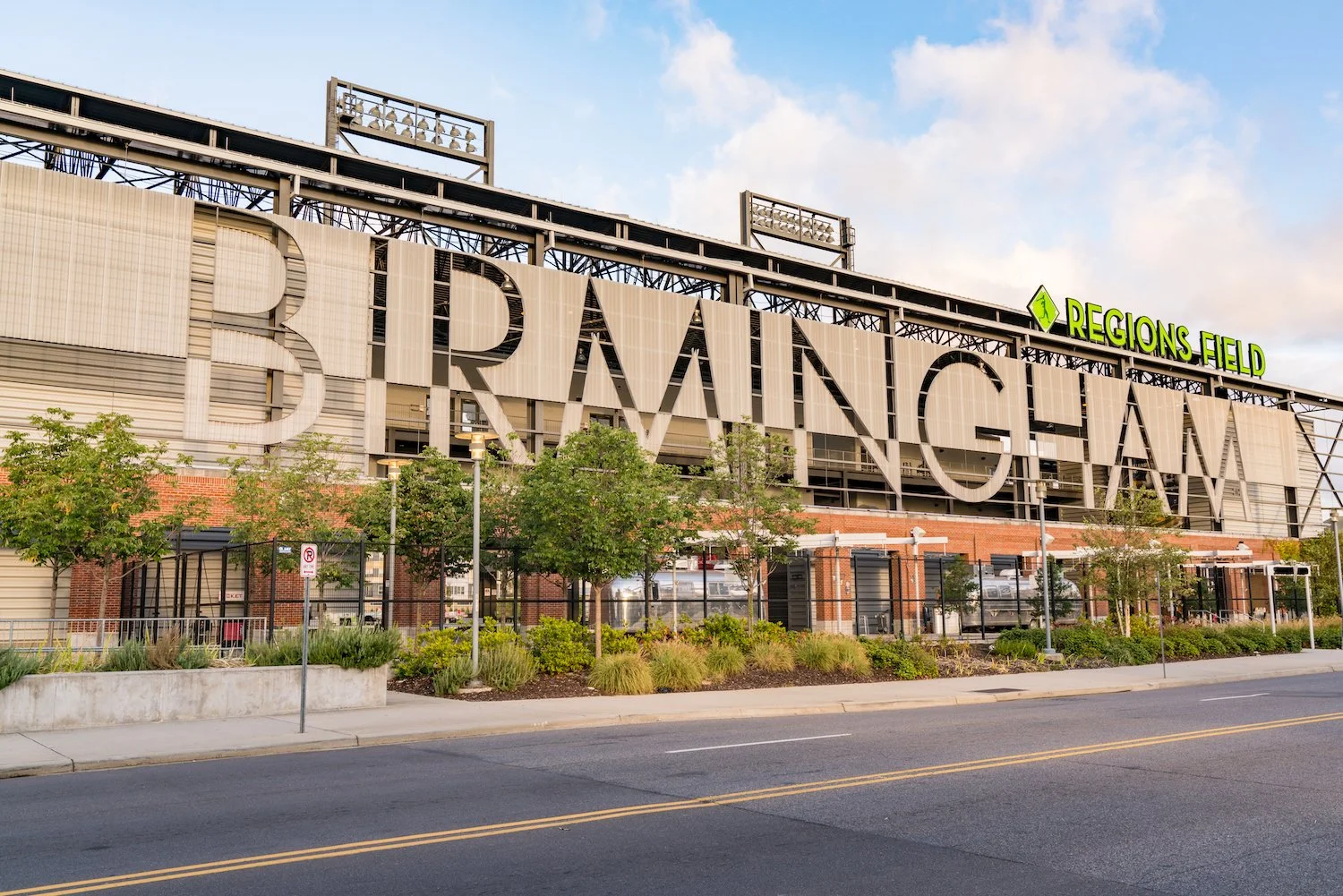 Exterior view of Regions Field, home of the Birmingham Barons, with large sign spelling 'BIRMINGHAM' on the building. Some trees, shrubs, a sidewalk, and street in the foreground, and a partly cloudy sky above.