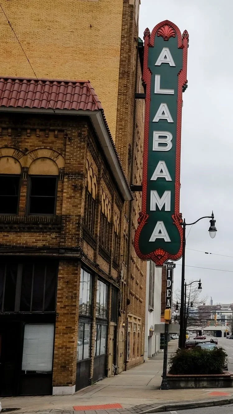 Vertical neon sign reading 'ALABAMA' attached to a brick building on a city sidewalk.